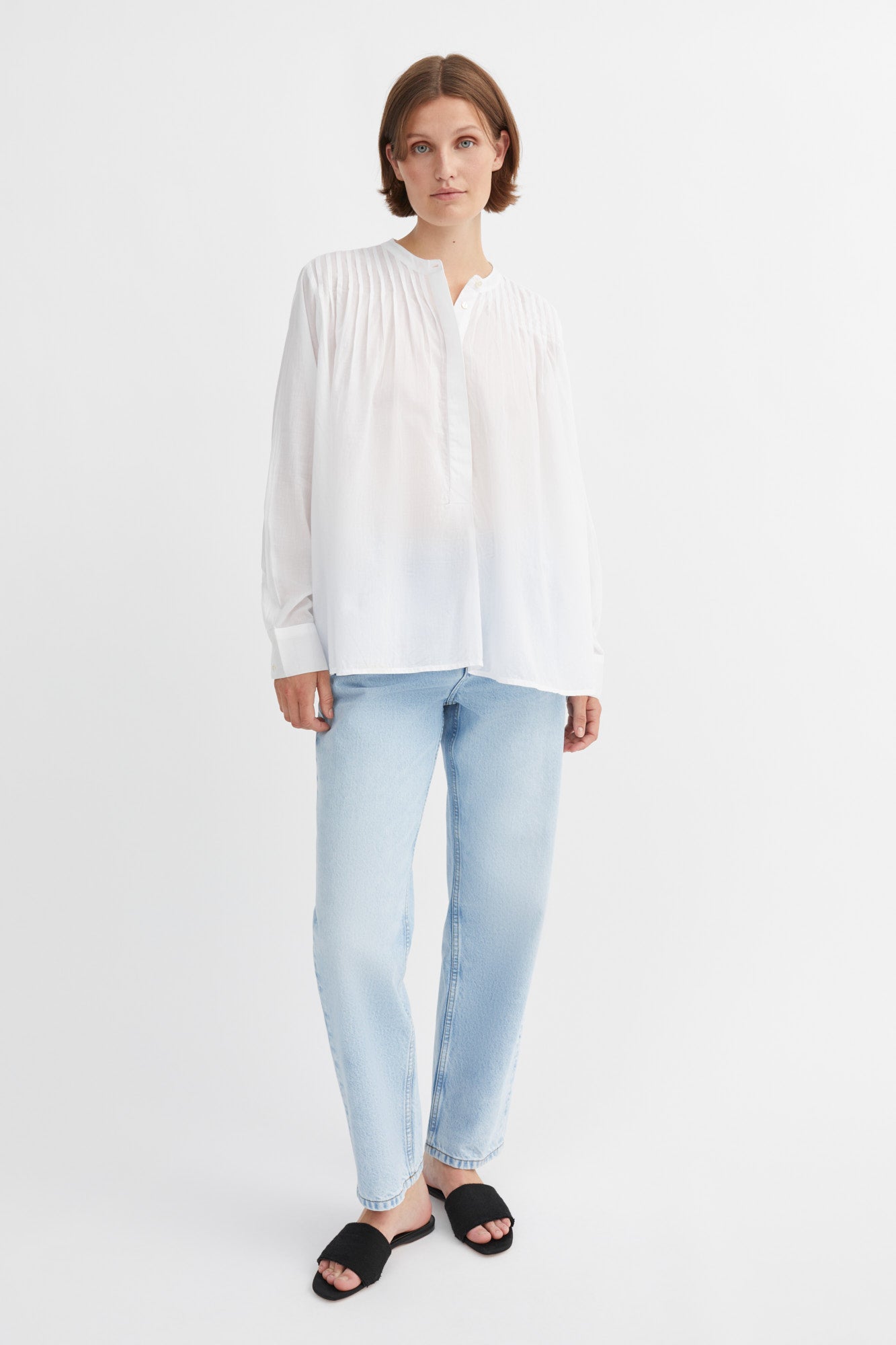 A woman modeling a white long-sleeve blouse with a gathered, pleated neckline, paired with light blue jeans and black slide sandals, against a plain white studio background.