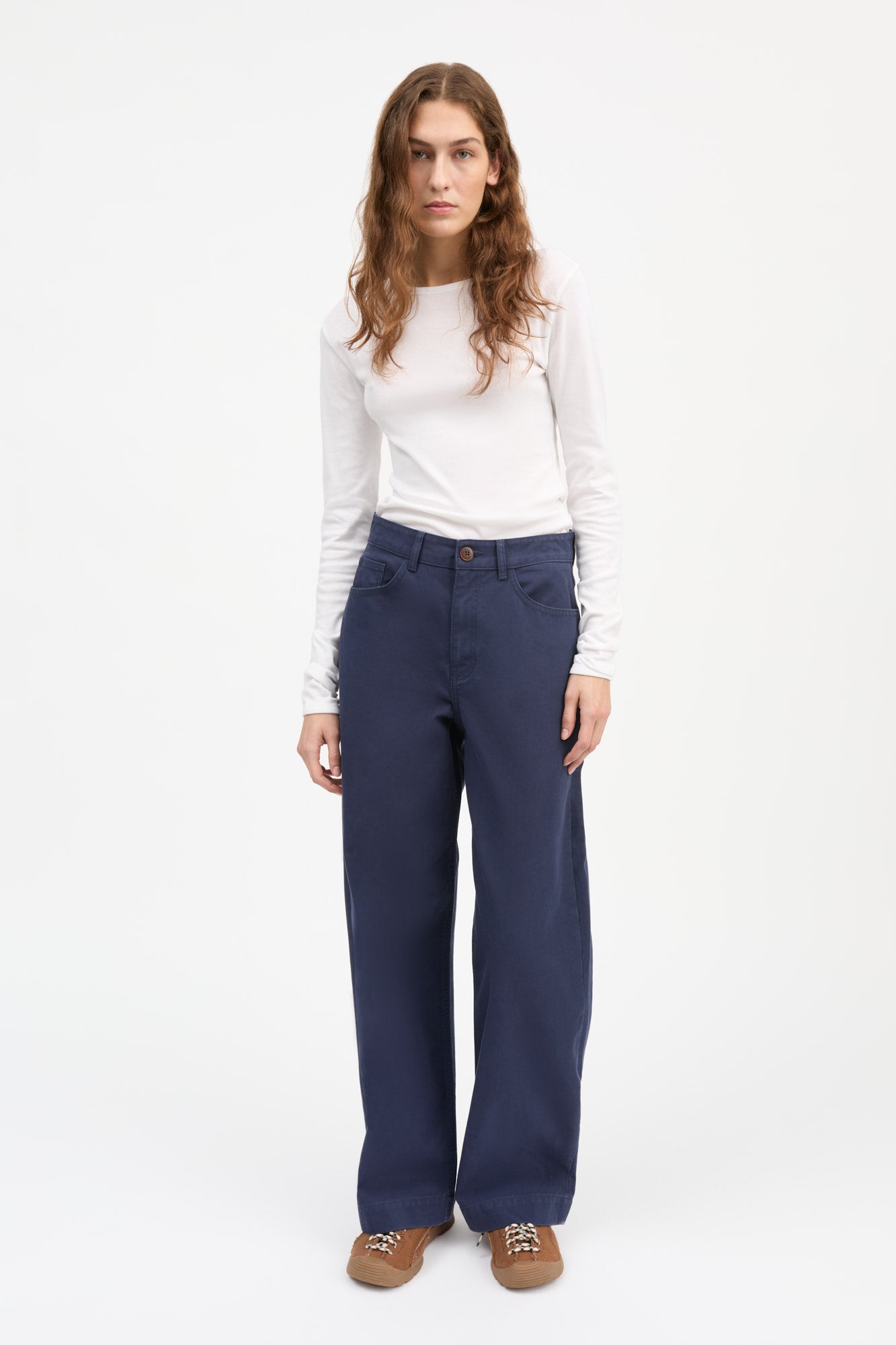 A woman modeling a pair of wide-leg navy blue pants with a white long-sleeve top and brown shoes, standing against a plain white studio background.