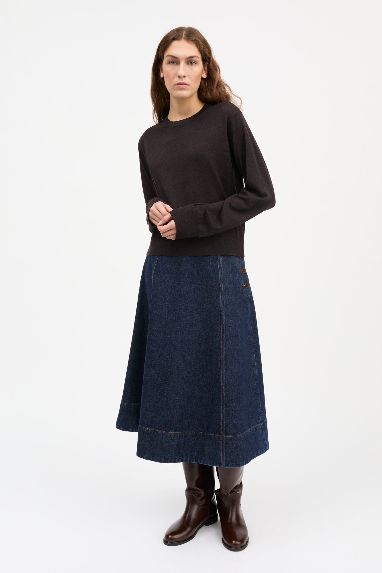 A woman modeling a dark brown long-sleeve crewneck sweater and a dark blue denim midi skirt, paired with dark brown leather ankle boots, against a plain white studio background.