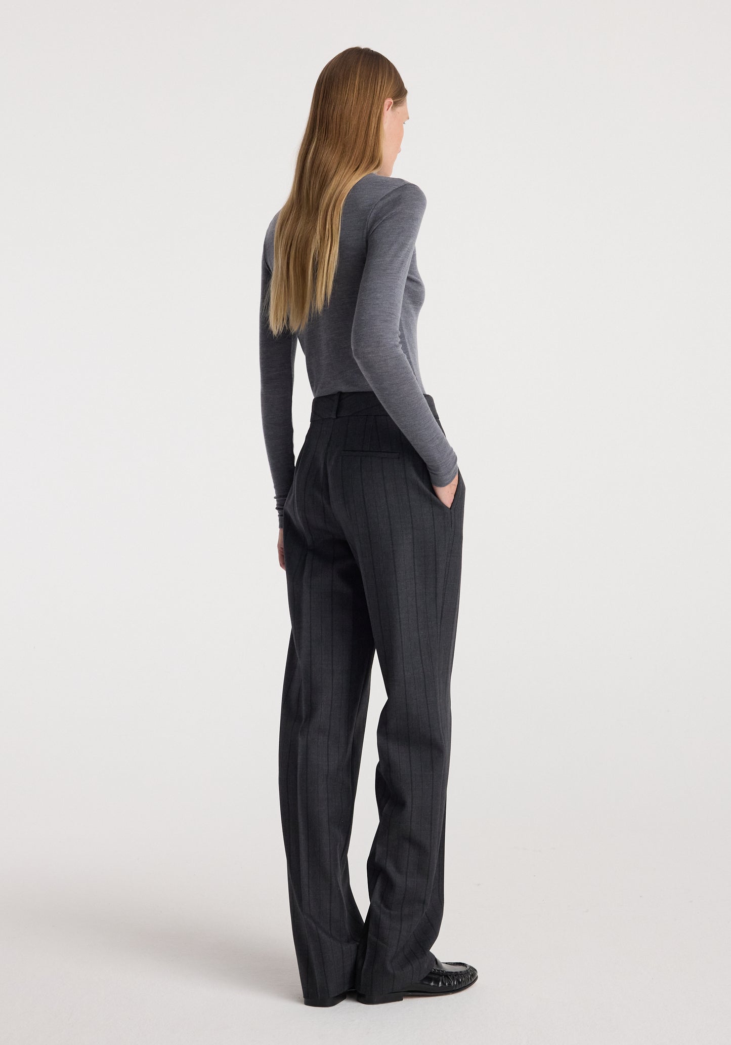 A woman is shown from the back wearing a gray long-sleeve top and dark charcoal gray pinstriped trousers, standing against a plain light gray studio background.