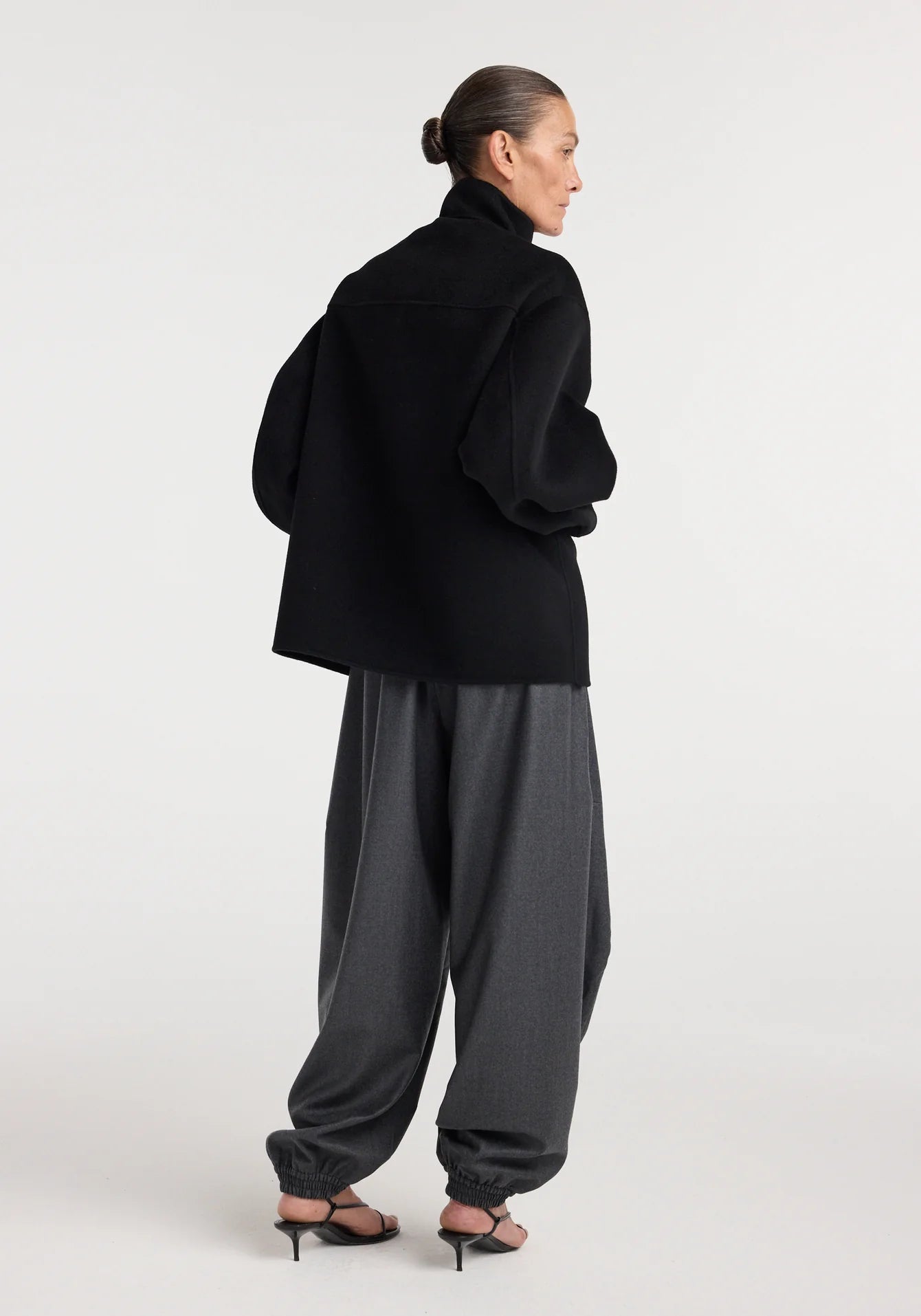 Person wearing a black outfit with a wide jacket and gray pants on a white background