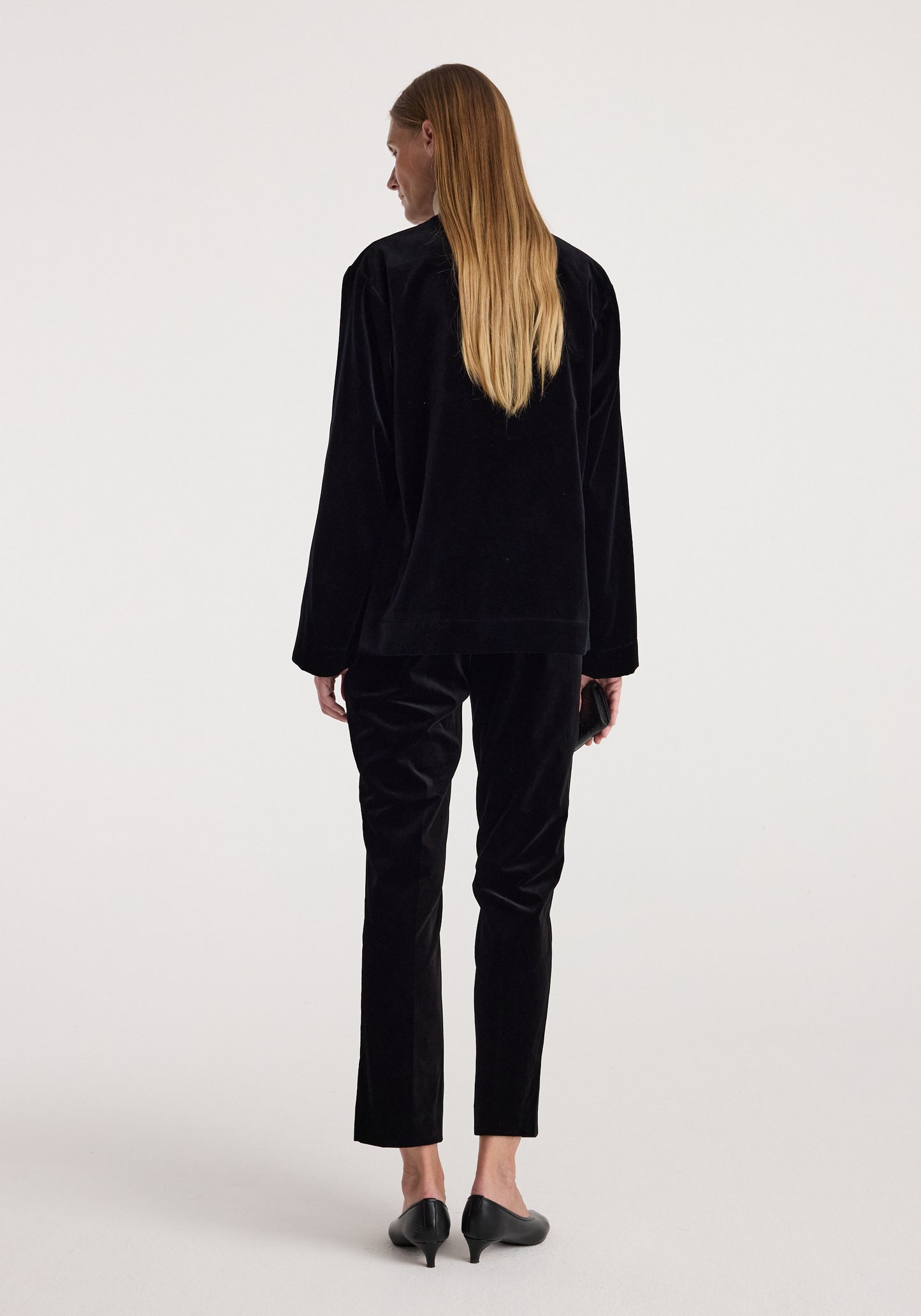 A woman with long blonde hair is shown from behind wearing a black velvet long-sleeve top and matching black velvet cropped trousers, paired with black pointed-toe heels, against a light gray studio background.