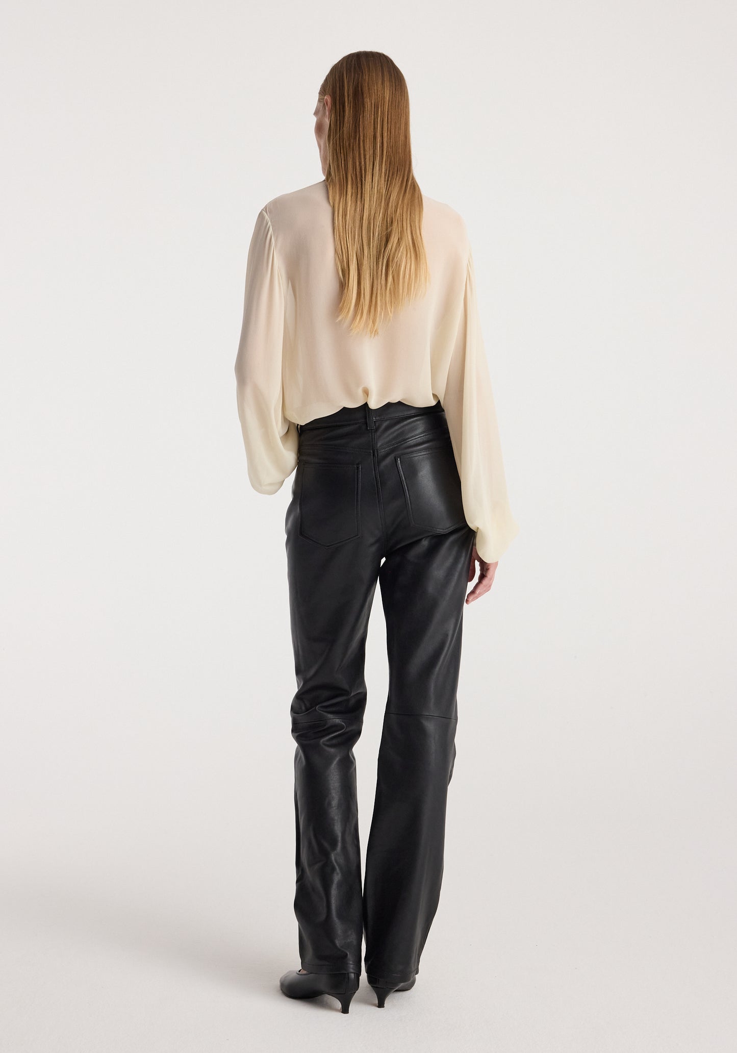 A woman viewed from behind wearing a cream-colored long-sleeve blouse and black leather pants against a plain light gray studio background.