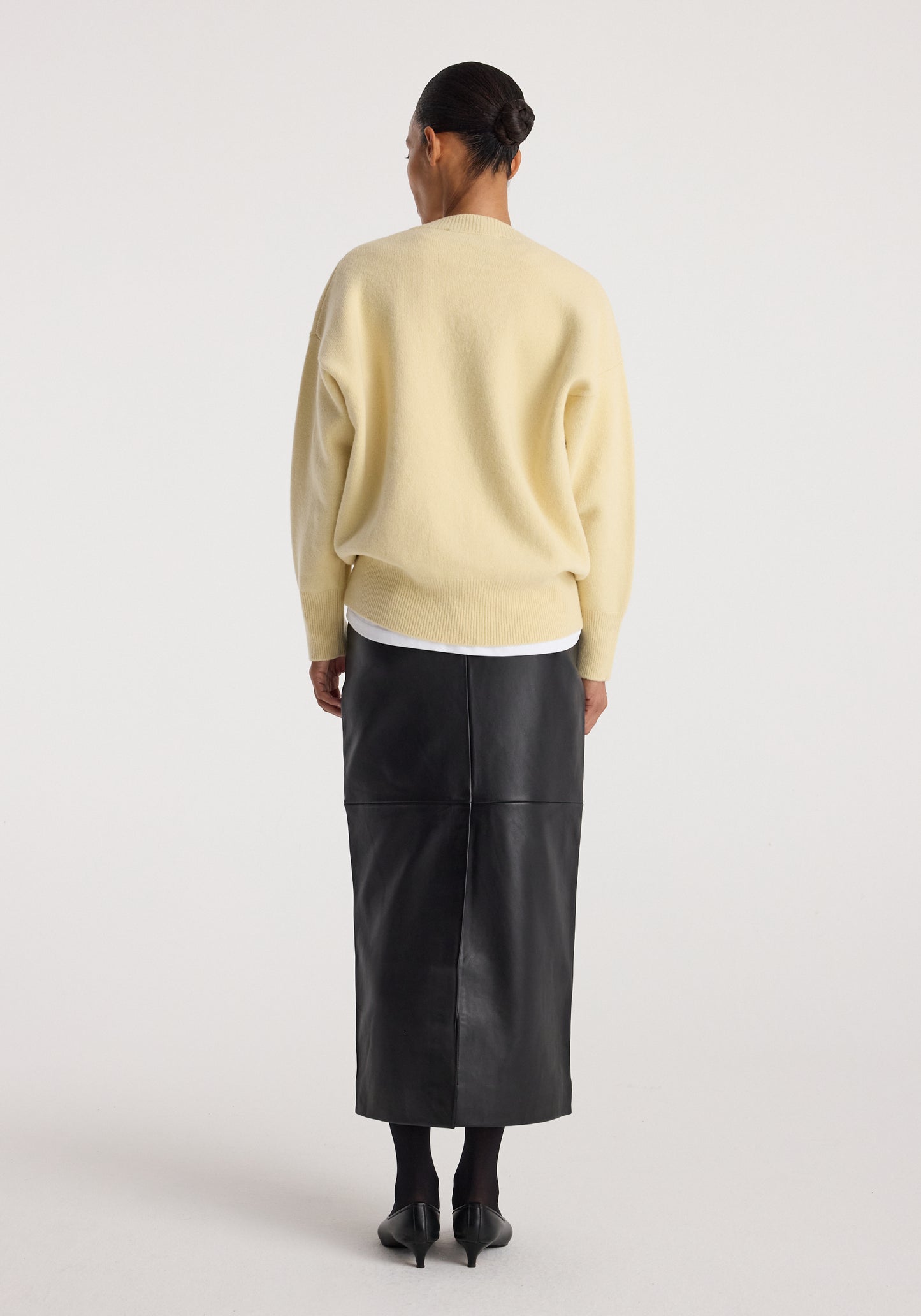 A model shown from the back wearing a pale yellow knit sweater, a black leather midi skirt, black tights, and black heels, against a plain light gray studio background.