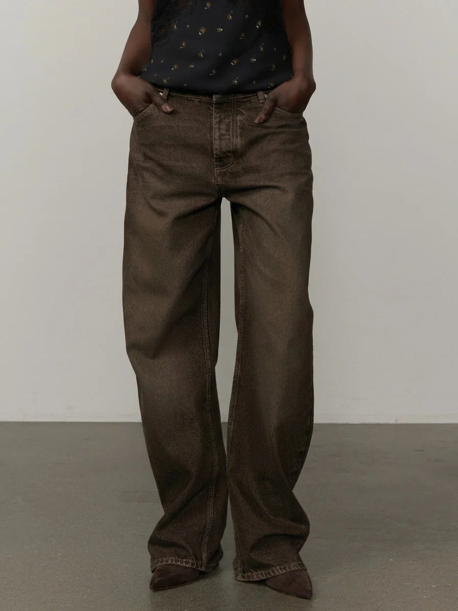 A person wearing dark brown wide-leg jeans with a black patterned top, standing against a plain light gray wall and concrete floor.