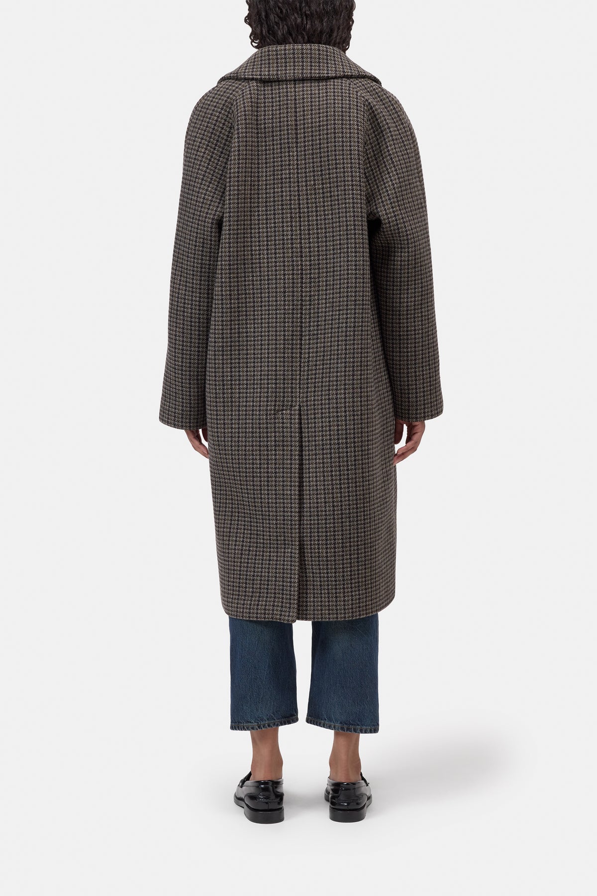 A person shown from the back wearing a long brown and black checkered coat, blue cropped jeans, and black shoes against a plain light gray studio background.