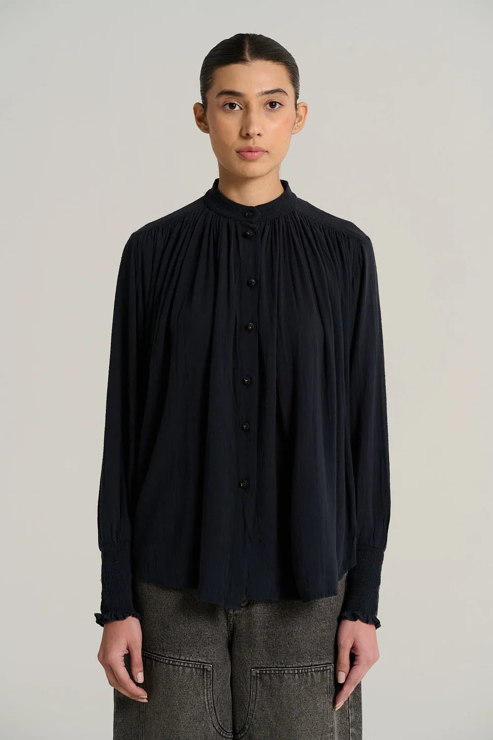 A model wearing a black long-sleeve blouse with a high gathered mock neck and front buttons, paired with gray trousers, against a plain light gray studio background.