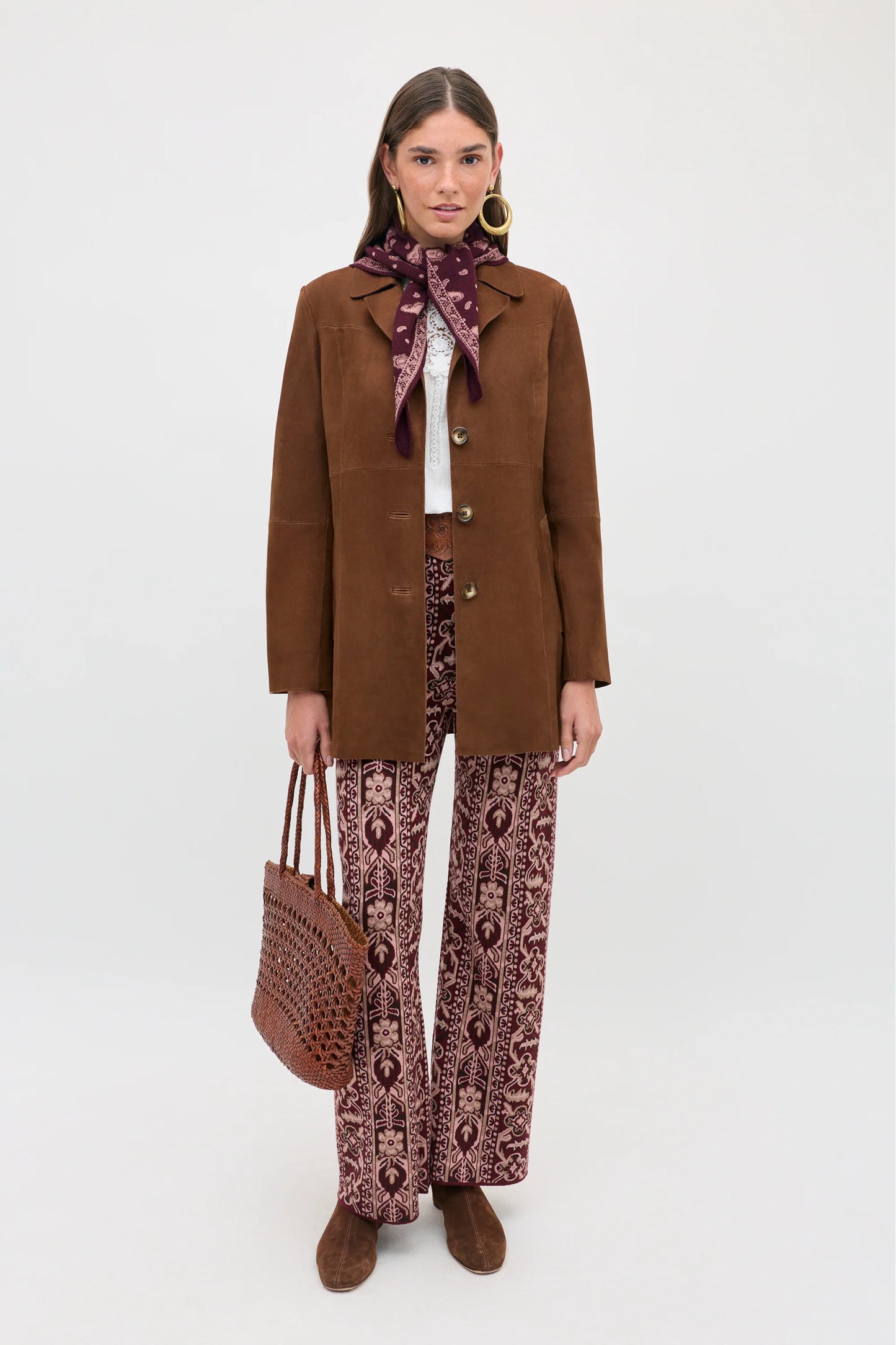 A model stands against a light gray studio background wearing a brown suede-like blazer, matching brown suede boots, and patterned brown and burgundy wide-leg trousers. She is styled with a burgundy and black patterned scarf, a white top, large gold hoop earrings, and a brown woven tote bag.