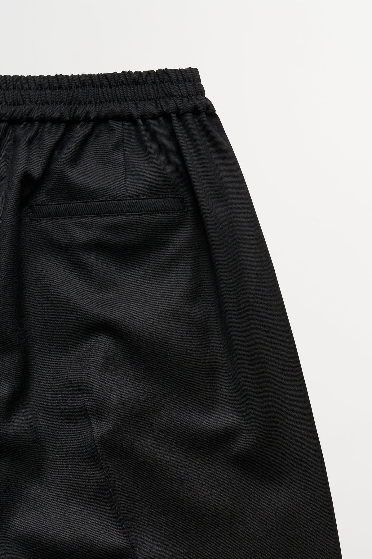 Close-up of black satin-like trousers showing the elastic waistband and a side pocket, photographed against a light gray background.