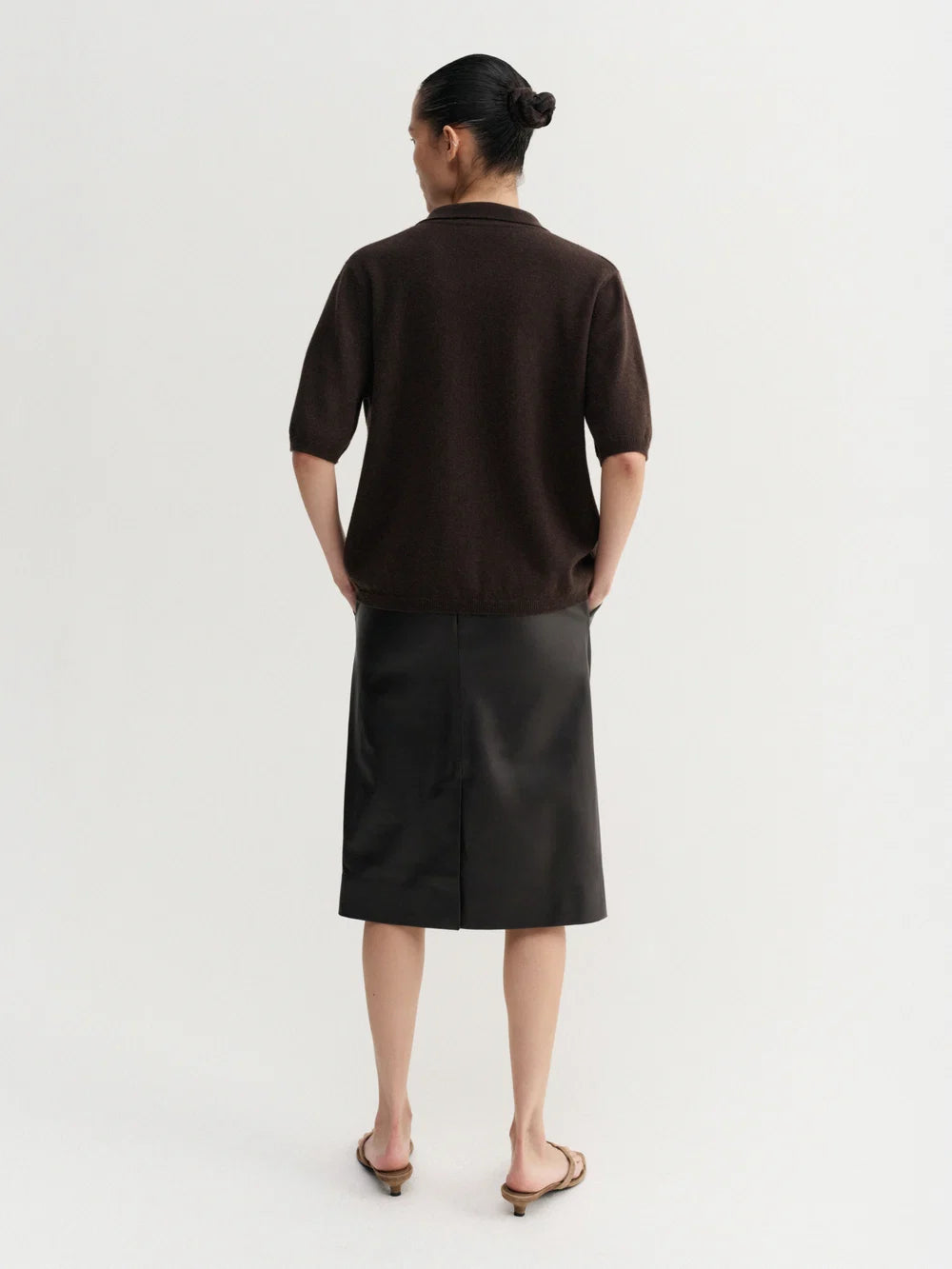 A model viewed from the back wearing a dark brown short-sleeve knit top and a black midi skirt, paired with brown flat sandals, against a plain white studio background.