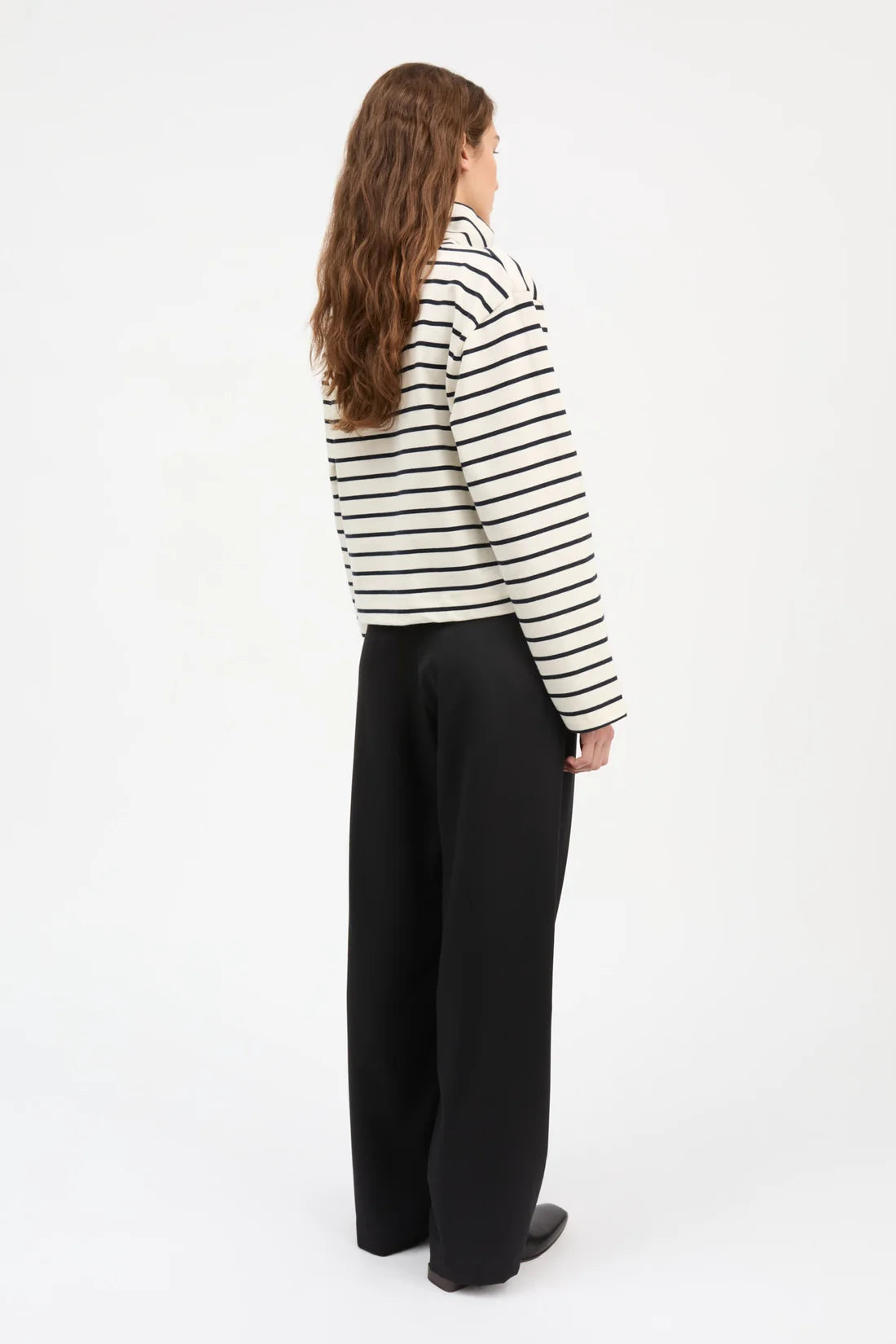 A woman shown from the back wearing a black and white horizontally striped long-sleeve top and black wide-leg trousers, standing against a plain white studio background.