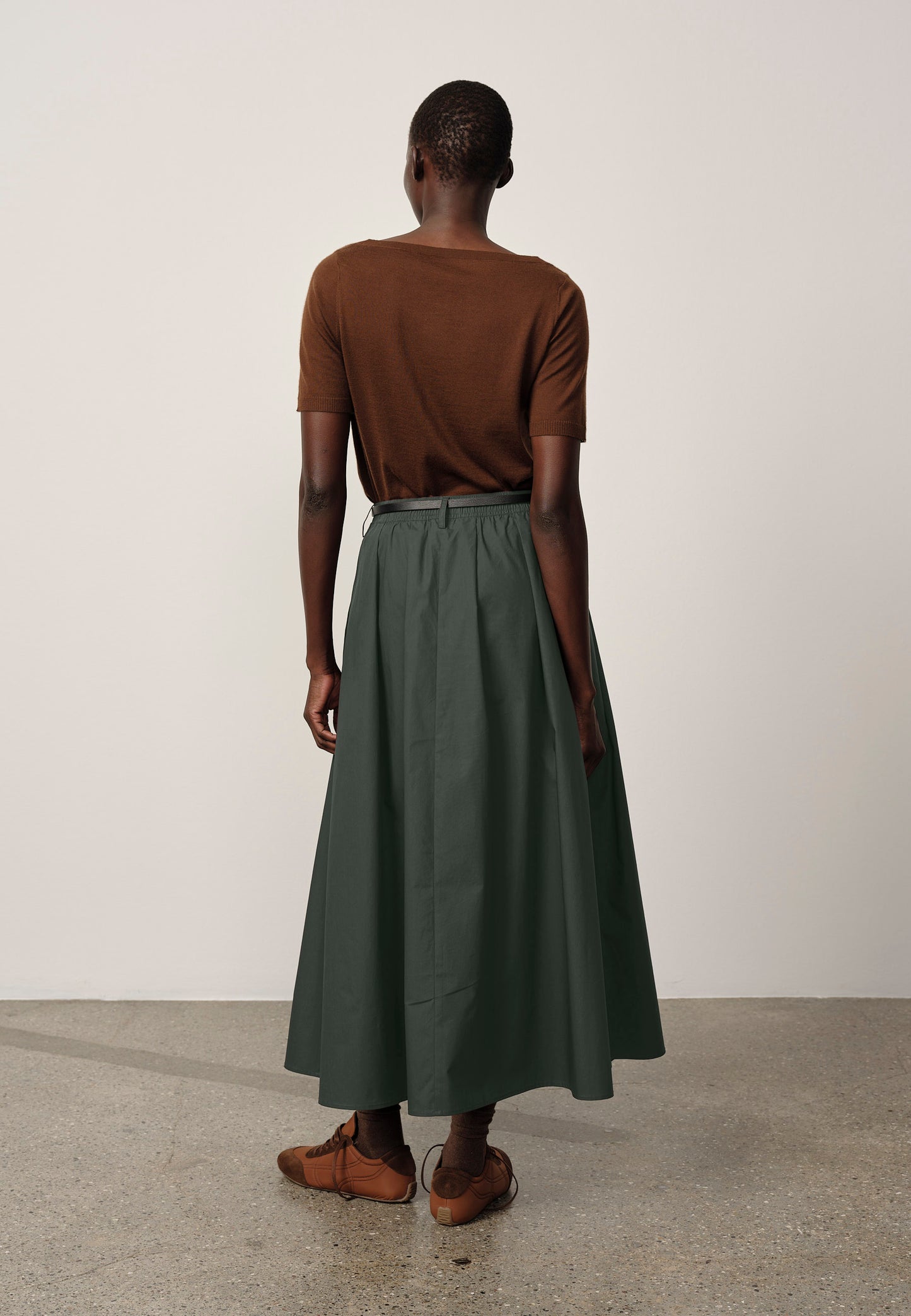 Person wearing a brown top and green skirt standing against a white wall.