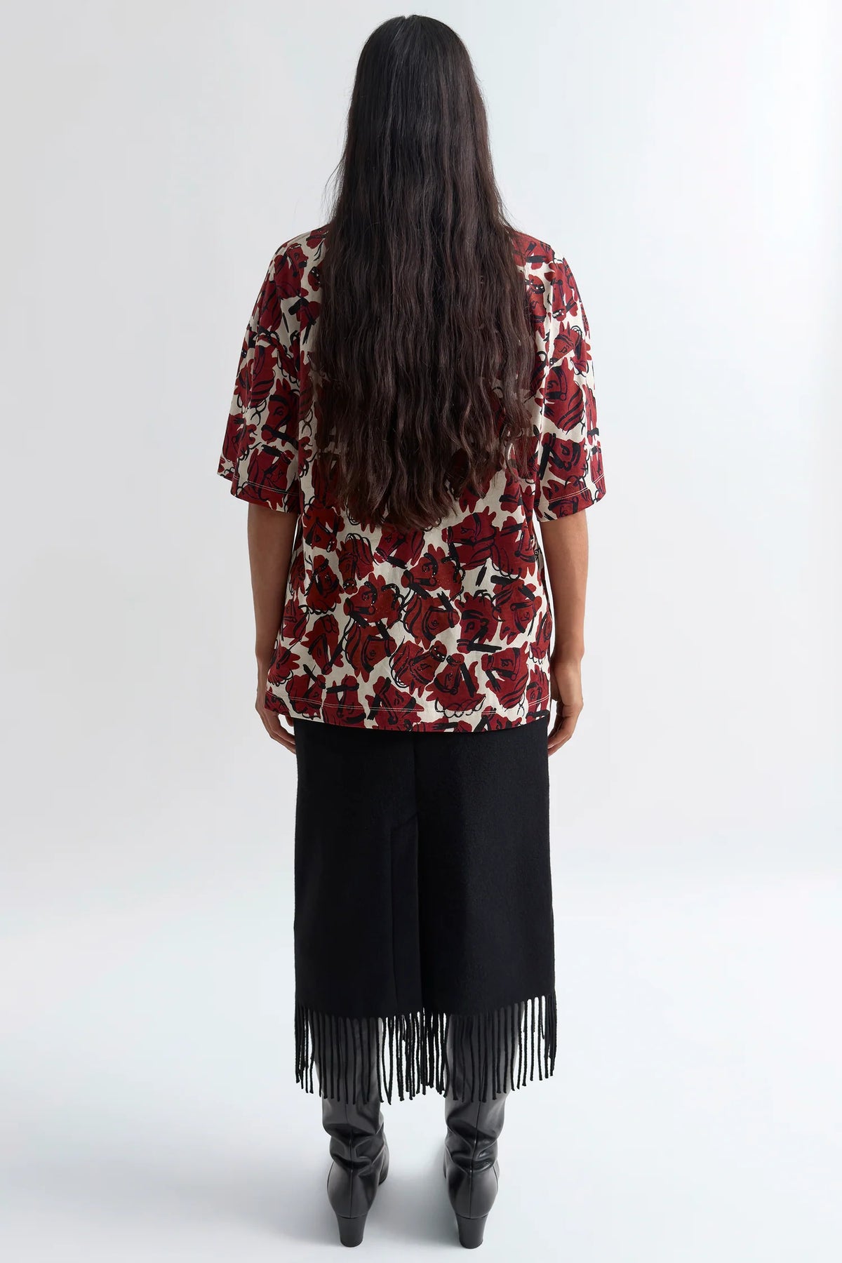 A woman viewed from behind wearing a short-sleeve top with a red and black floral pattern on a cream background, paired with a black skirt featuring fringe at the hem and black pointed boots, against a plain light gray studio background.