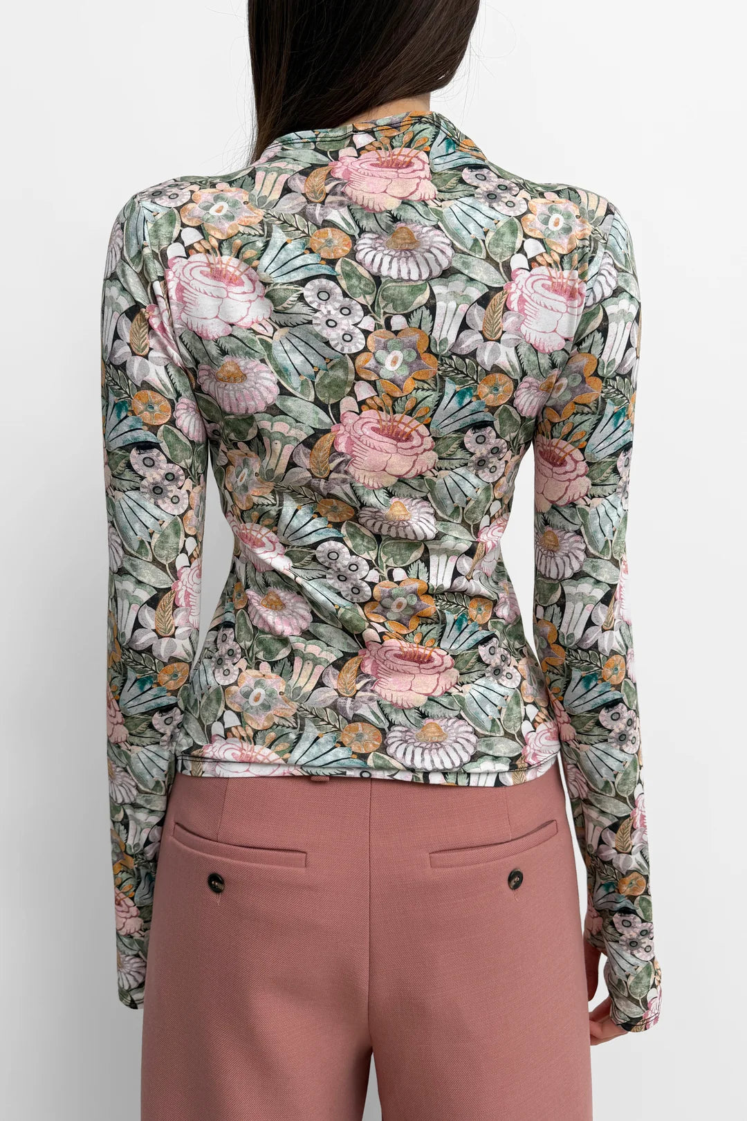 A woman shown from the back wearing a fitted long-sleeve top with an all-over floral print in muted green, pink, beige, and orange tones, paired with dusty rose trousers against a light gray studio background.