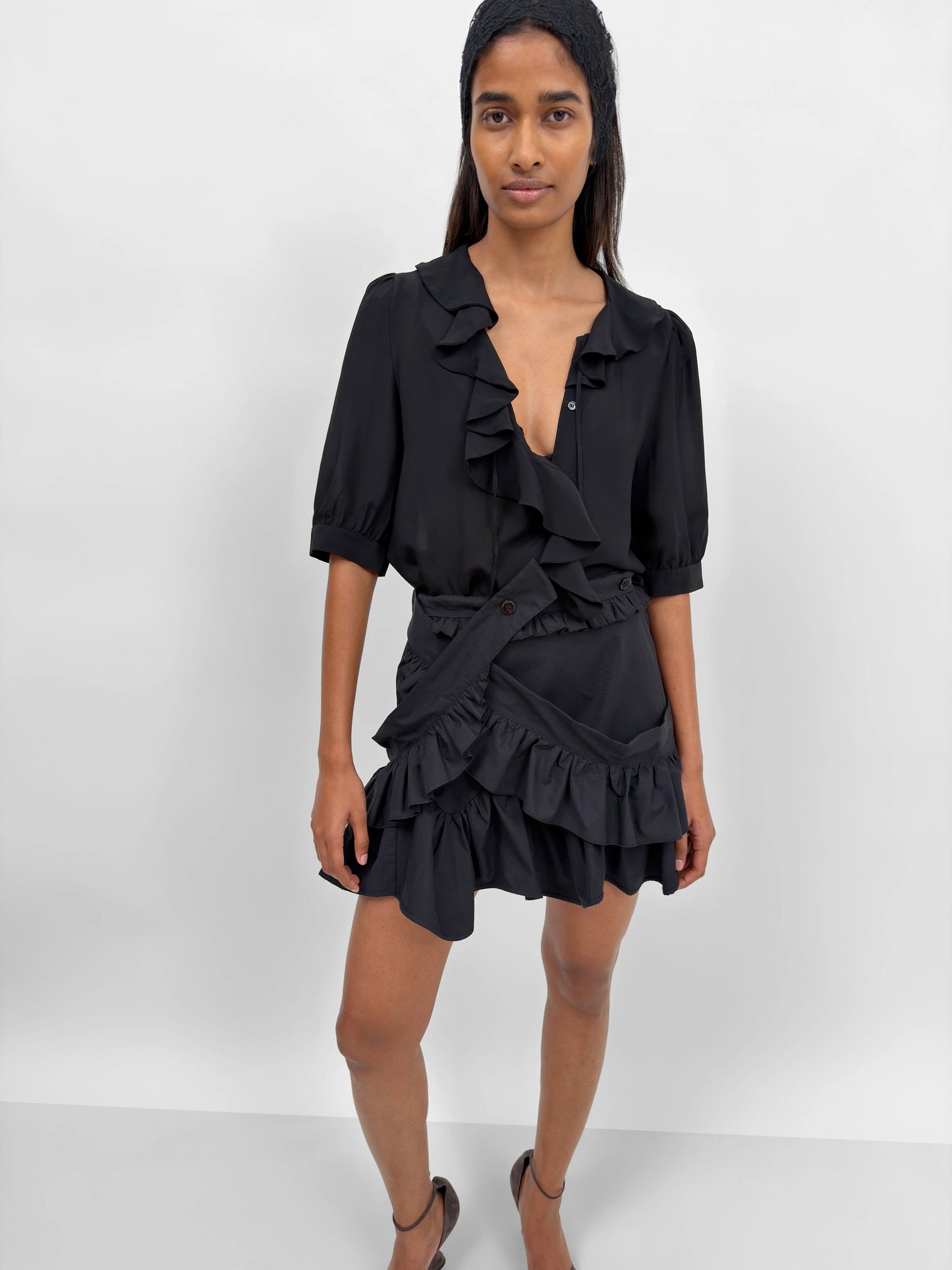 A woman modeling a black ruffled mini dress with puff sleeves, a deep V-neckline, and a matching ruffled belt, set against a plain light gray studio background.