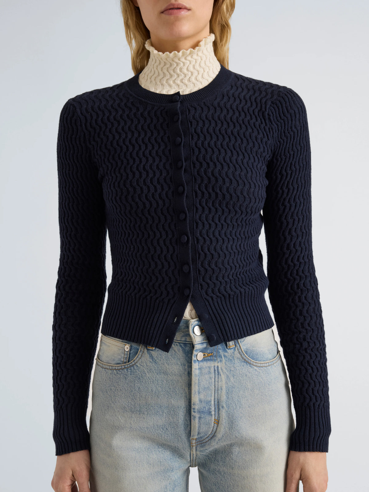 A model wearing a dark navy blue cable-knit cardigan over a cream turtleneck and light-wash blue jeans, shown from the neck down against a light gray studio background.