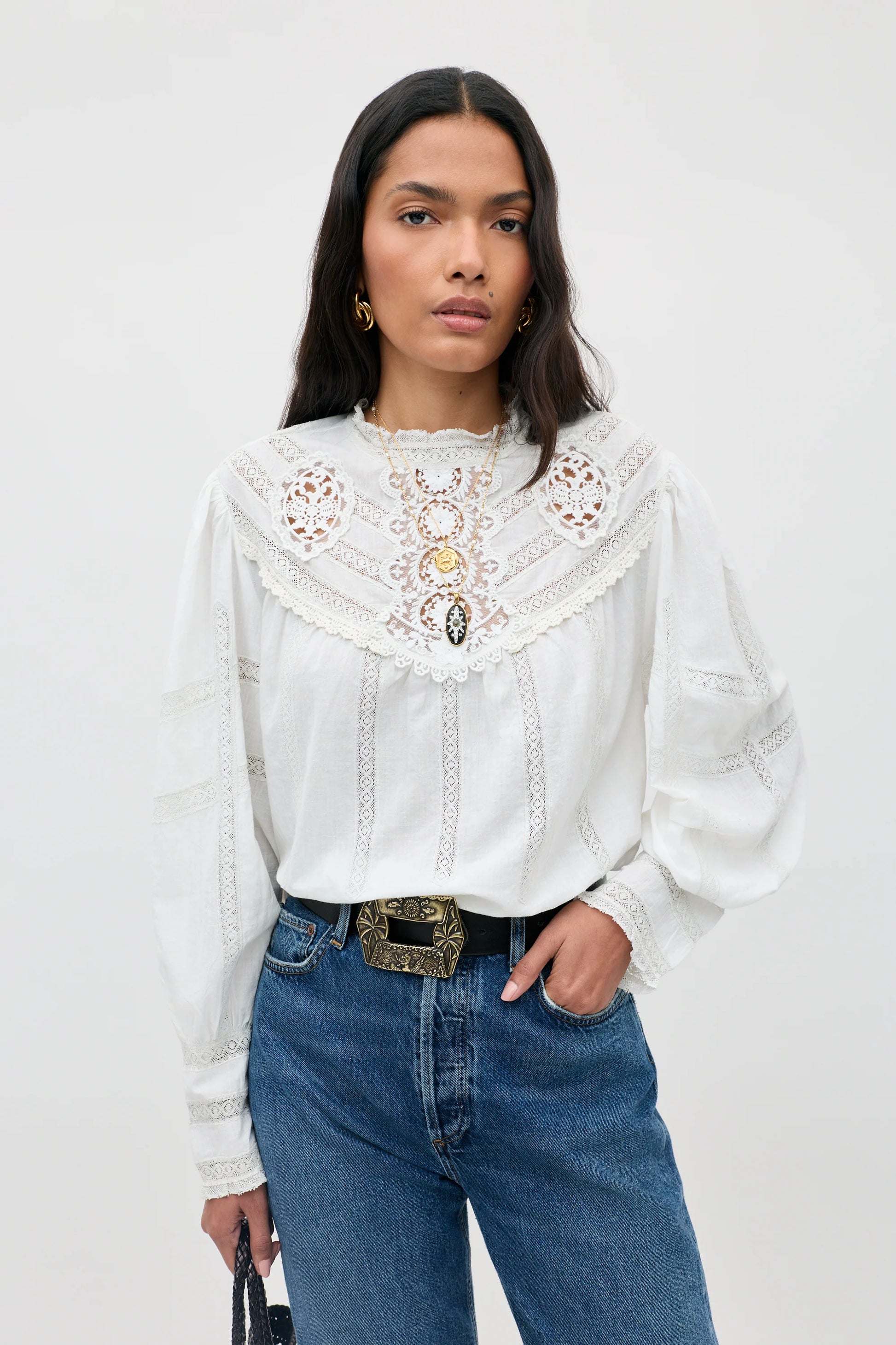 A woman modeling a white long-sleeve blouse with intricate lace and embroidery on the yoke, paired with blue jeans and a decorative belt, against a plain light gray studio background.