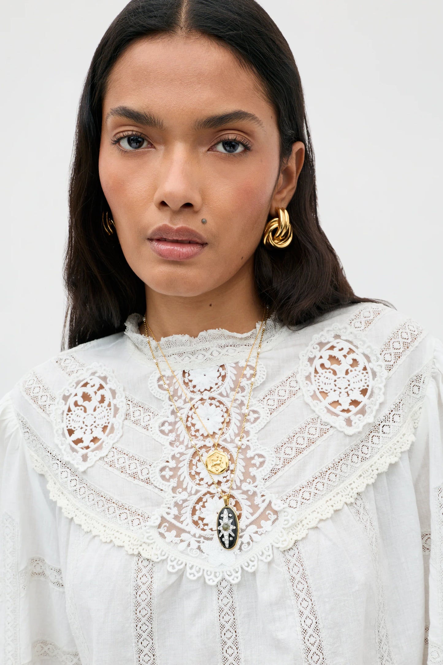 A close-up of a model wearing a white lace blouse with intricate embroidery and a large decorative collar, accessorized with gold jewelry against a light gray background.
