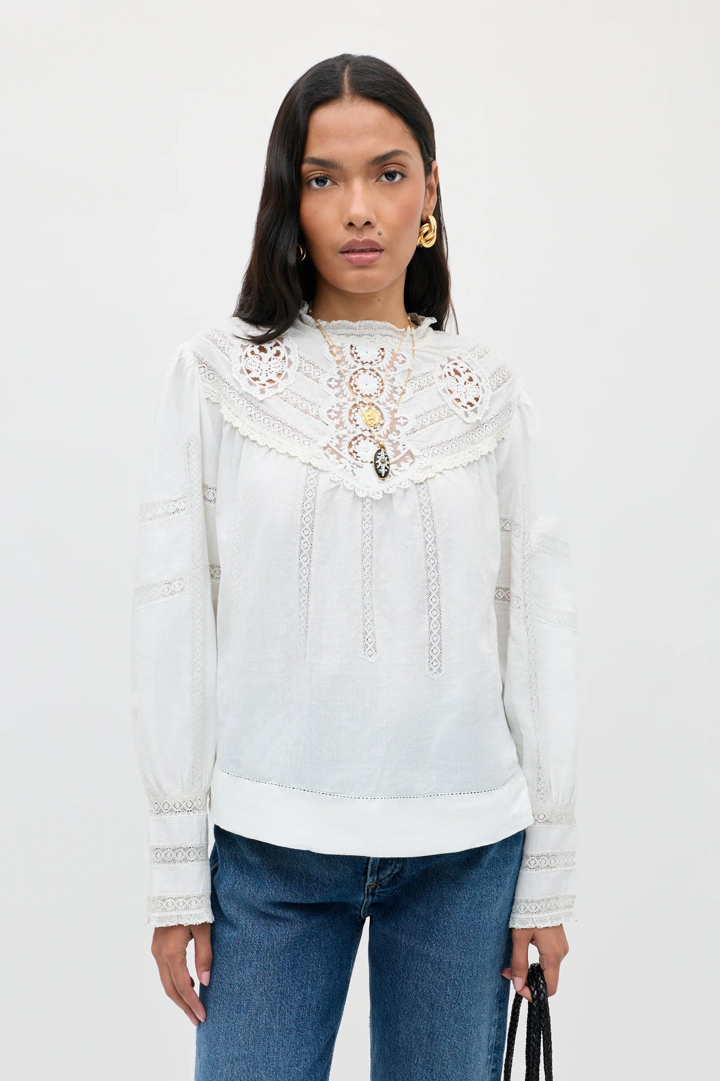 A woman modeling a white long-sleeve blouse with intricate lace and embroidery on the yoke and cuffs, paired with blue jeans against a plain white studio background.