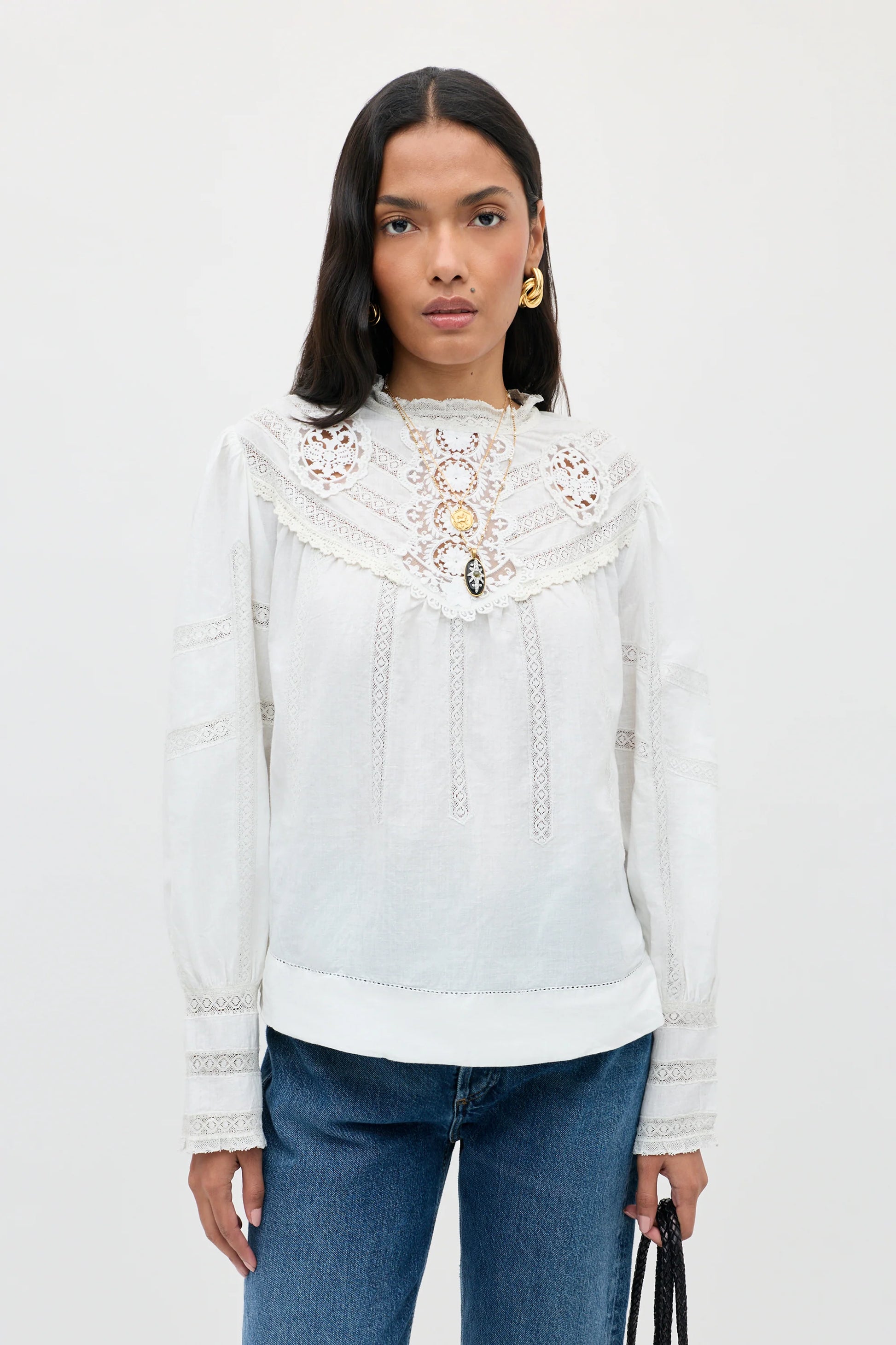A woman modeling a white long-sleeve blouse with intricate lace and embroidery on the yoke and cuffs, paired with blue jeans against a plain white studio background.