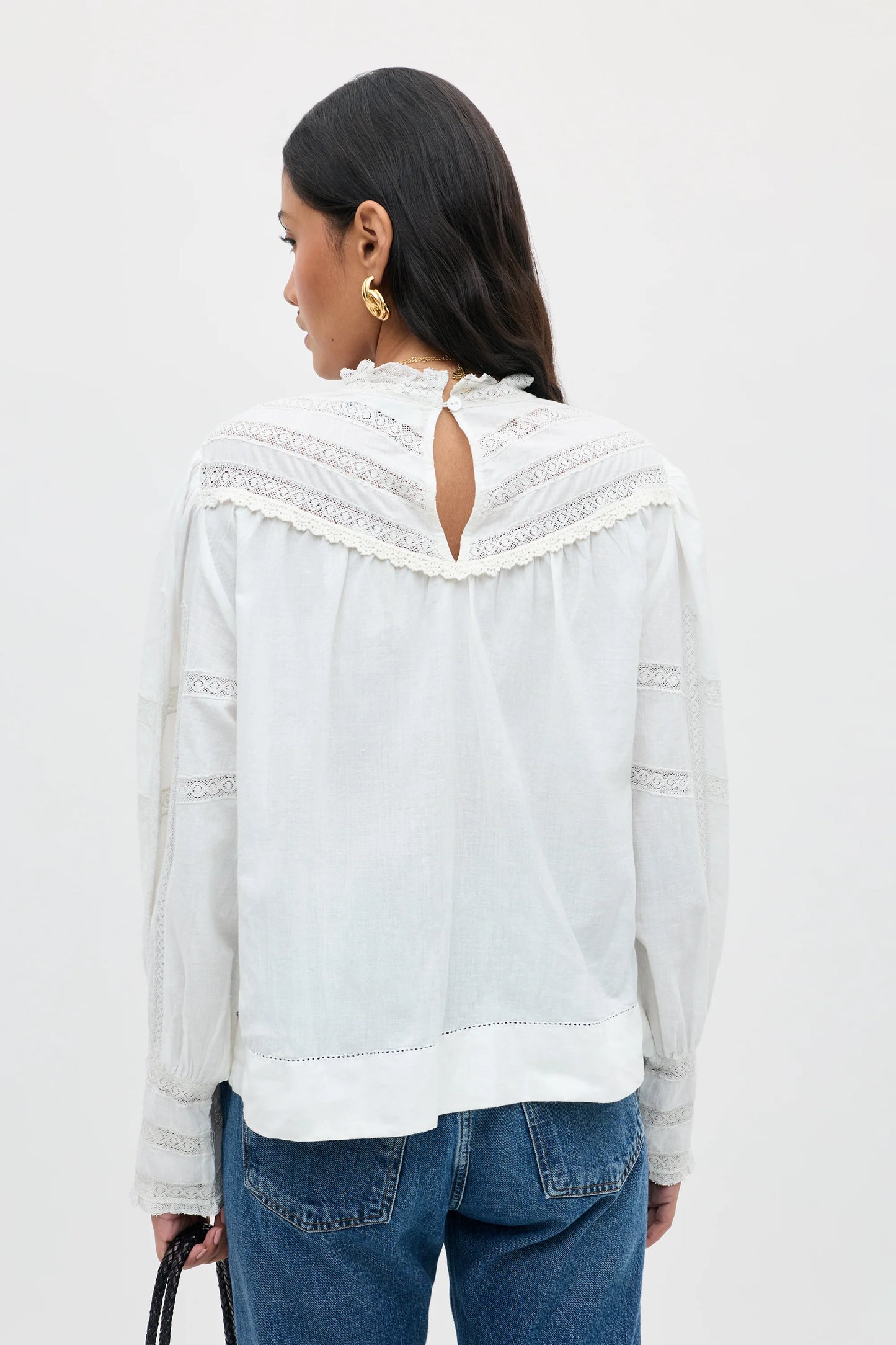A woman shown from the back wearing a white long-sleeve blouse with lace trim and a keyhole cutout at the neckline, paired with blue jeans against a plain light background.