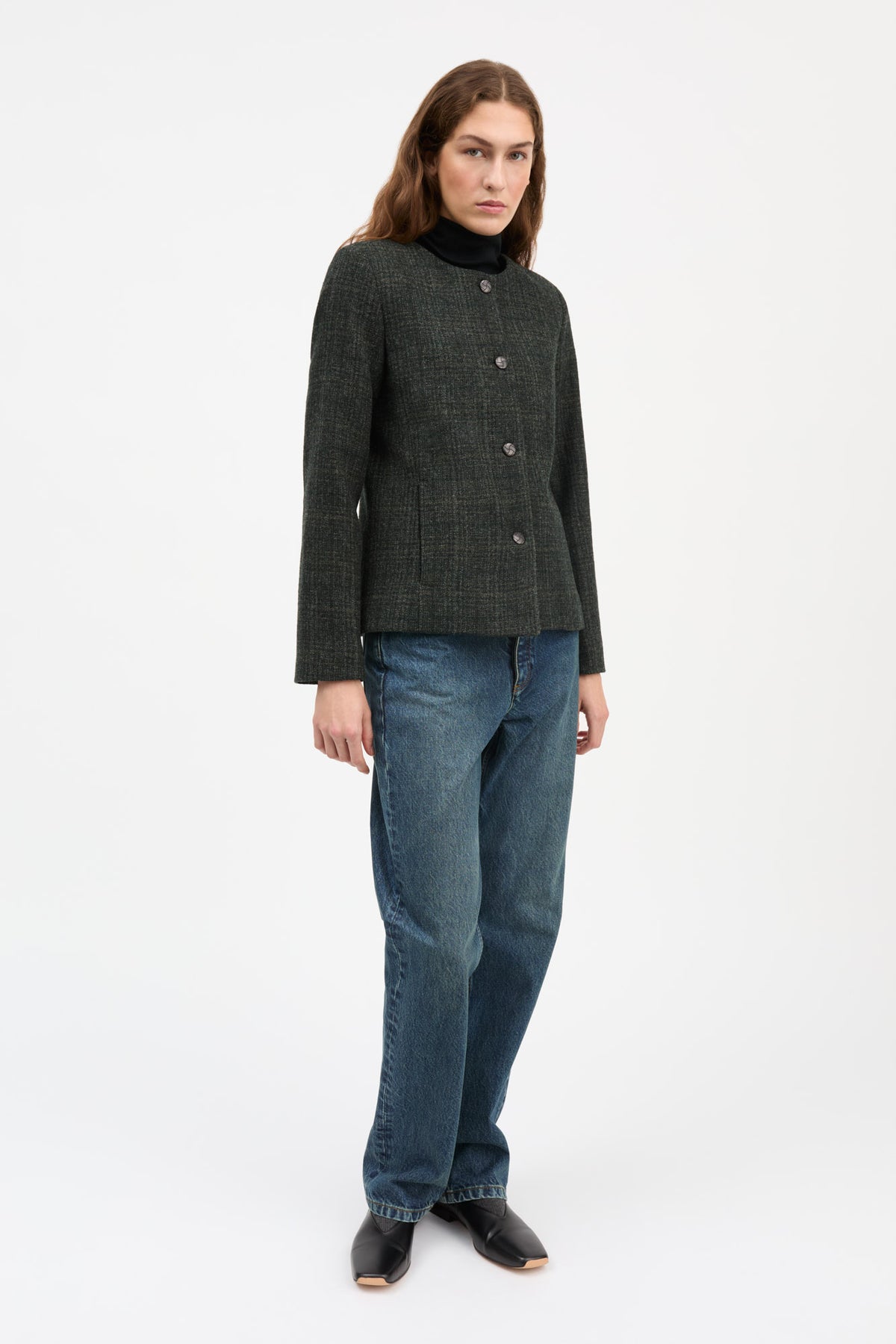 A woman modeling a dark charcoal tweed jacket with a stand collar and front buttons, paired with blue jeans and black shoes, against a plain white studio background.