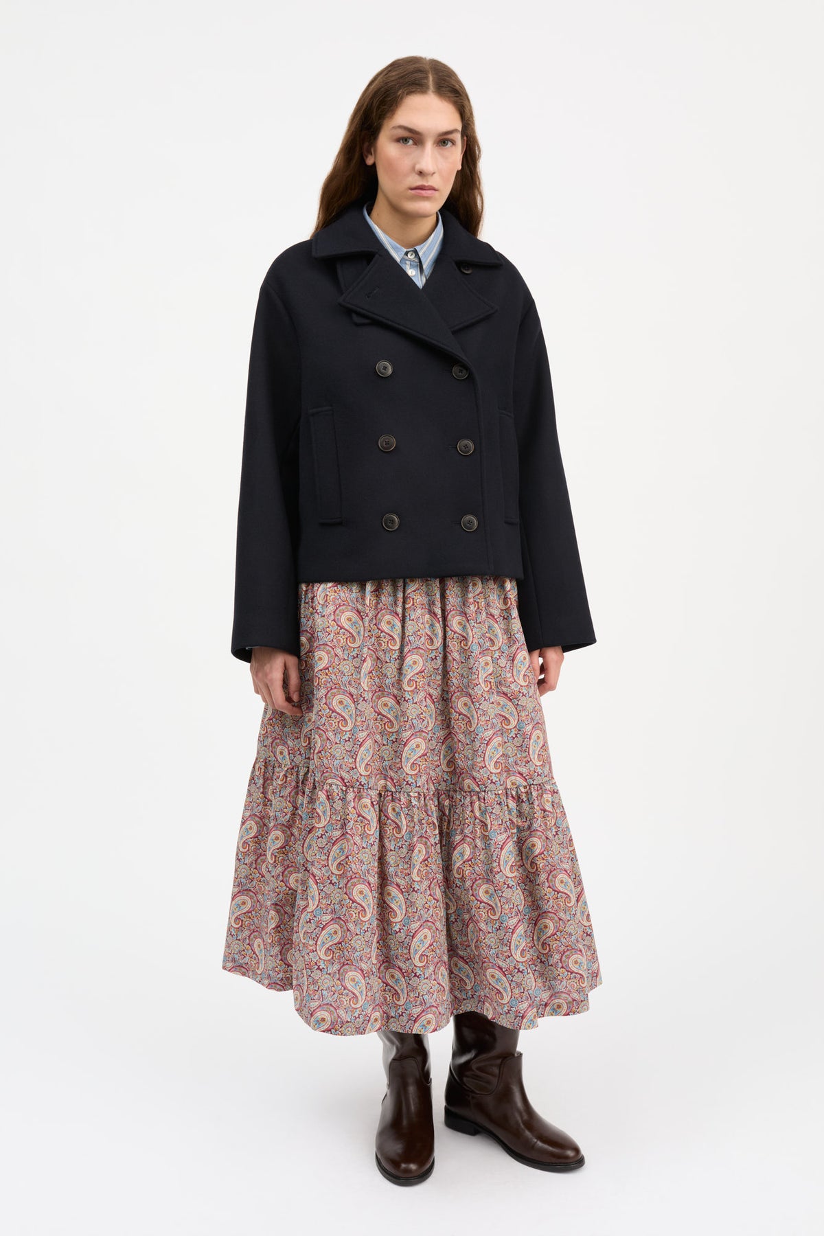 A model stands against a plain white studio background wearing a dark navy double-breasted coat over a light blue collared shirt, paired with a long tiered skirt in a pink and orange paisley pattern, and dark brown ankle boots.
