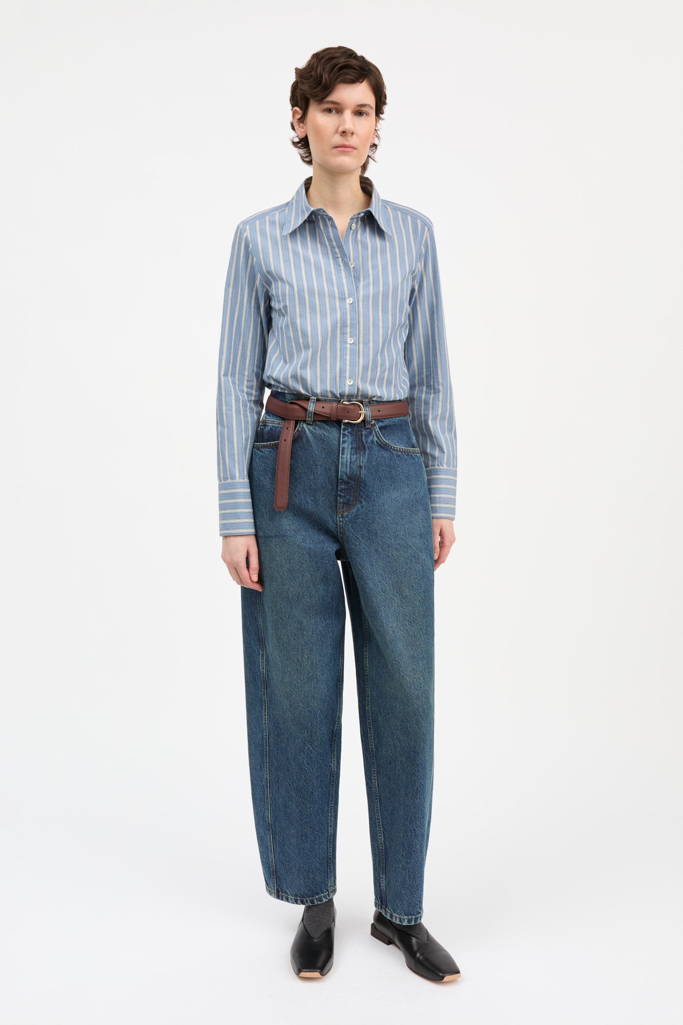 A model stands against a plain white studio background wearing a light blue and white vertically striped button-down shirt tucked into dark blue wide-leg jeans, styled with a brown leather belt and black leather shoes.