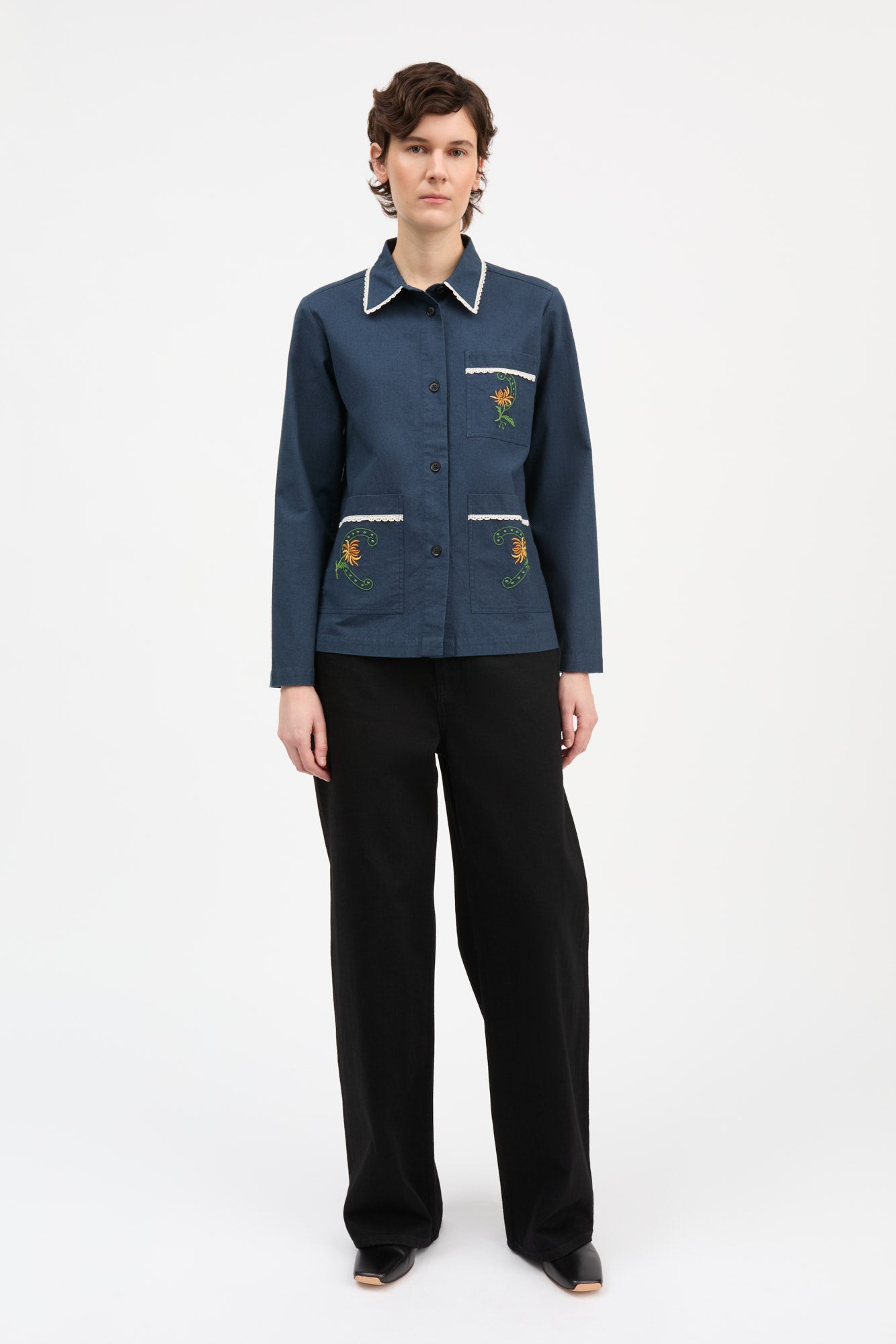 A model stands against a plain white studio background wearing a dark blue button-front jacket with white piping and embroidered floral details on the chest and lower front, paired with black wide-leg trousers.
