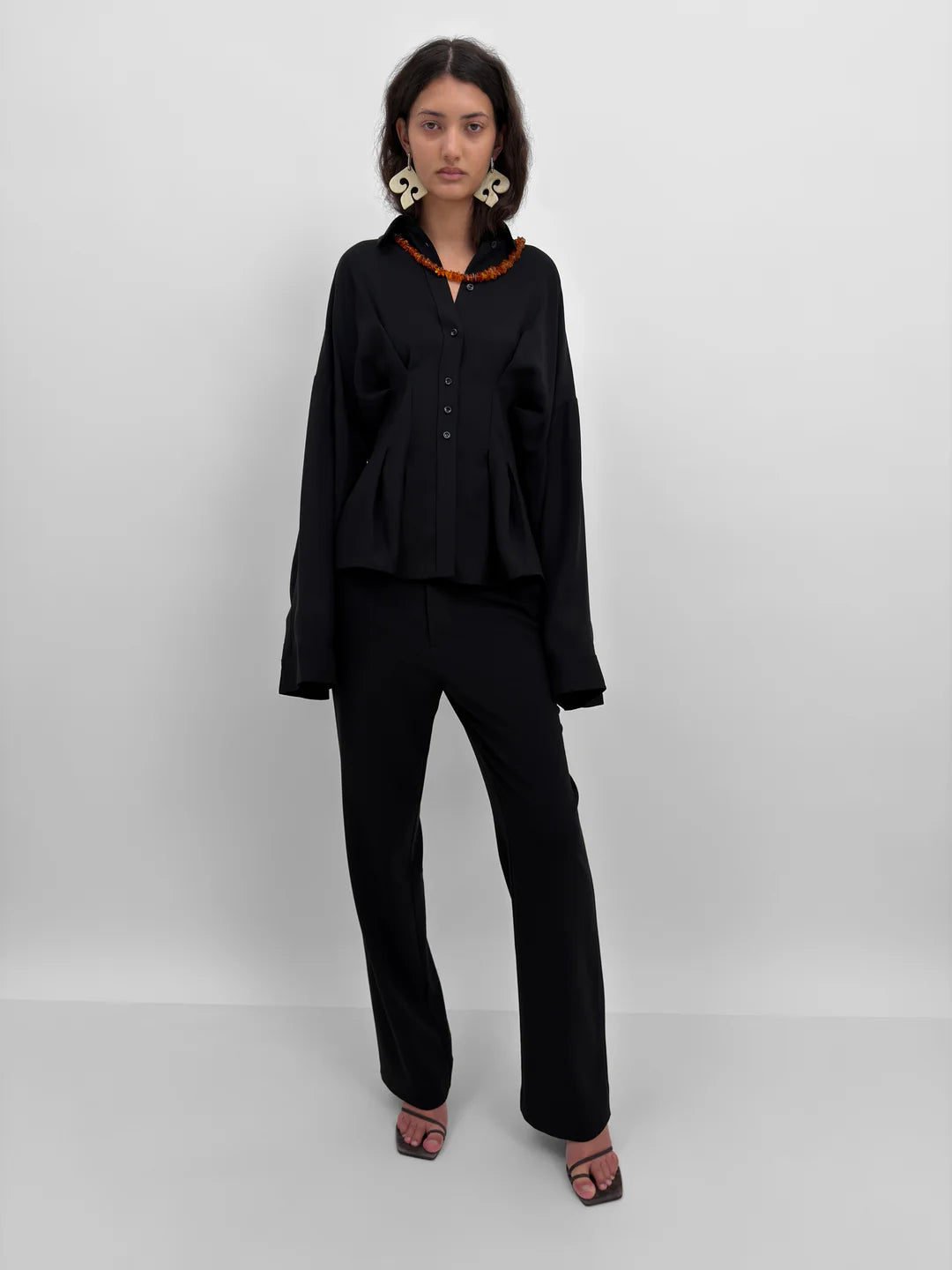 A model stands against a light gray studio background wearing a black long-sleeve button-front top with a peplum hem, black wide-leg pants, and black strappy heels. She is also wearing large white and black statement earrings and a chunky orange necklace.