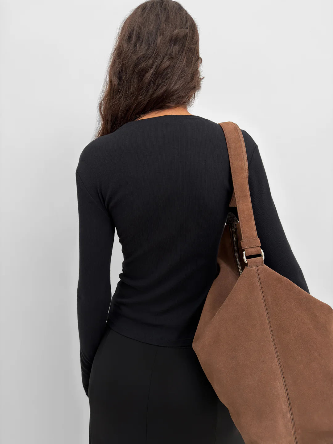 A woman shown from behind wearing a black long-sleeve top and skirt, carrying a large brown suede shoulder bag against a plain light gray background.