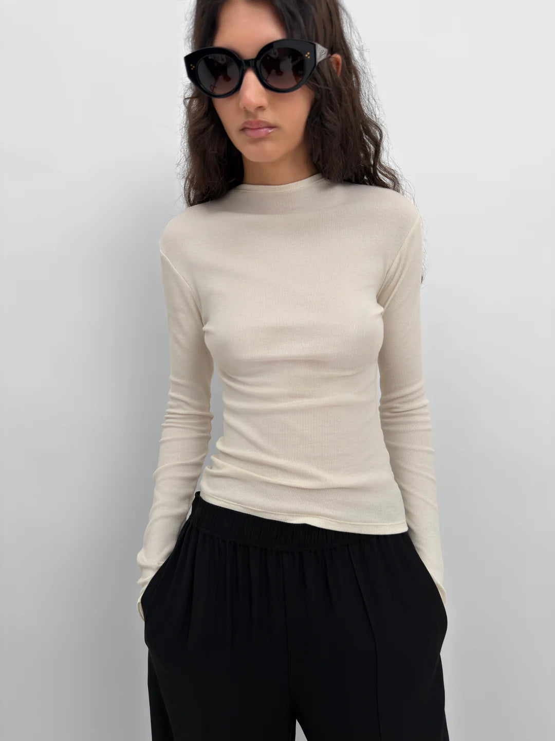 A woman wearing a fitted, long-sleeve cream-colored mock neck top and black pants, styled with black sunglasses against a plain light gray background.