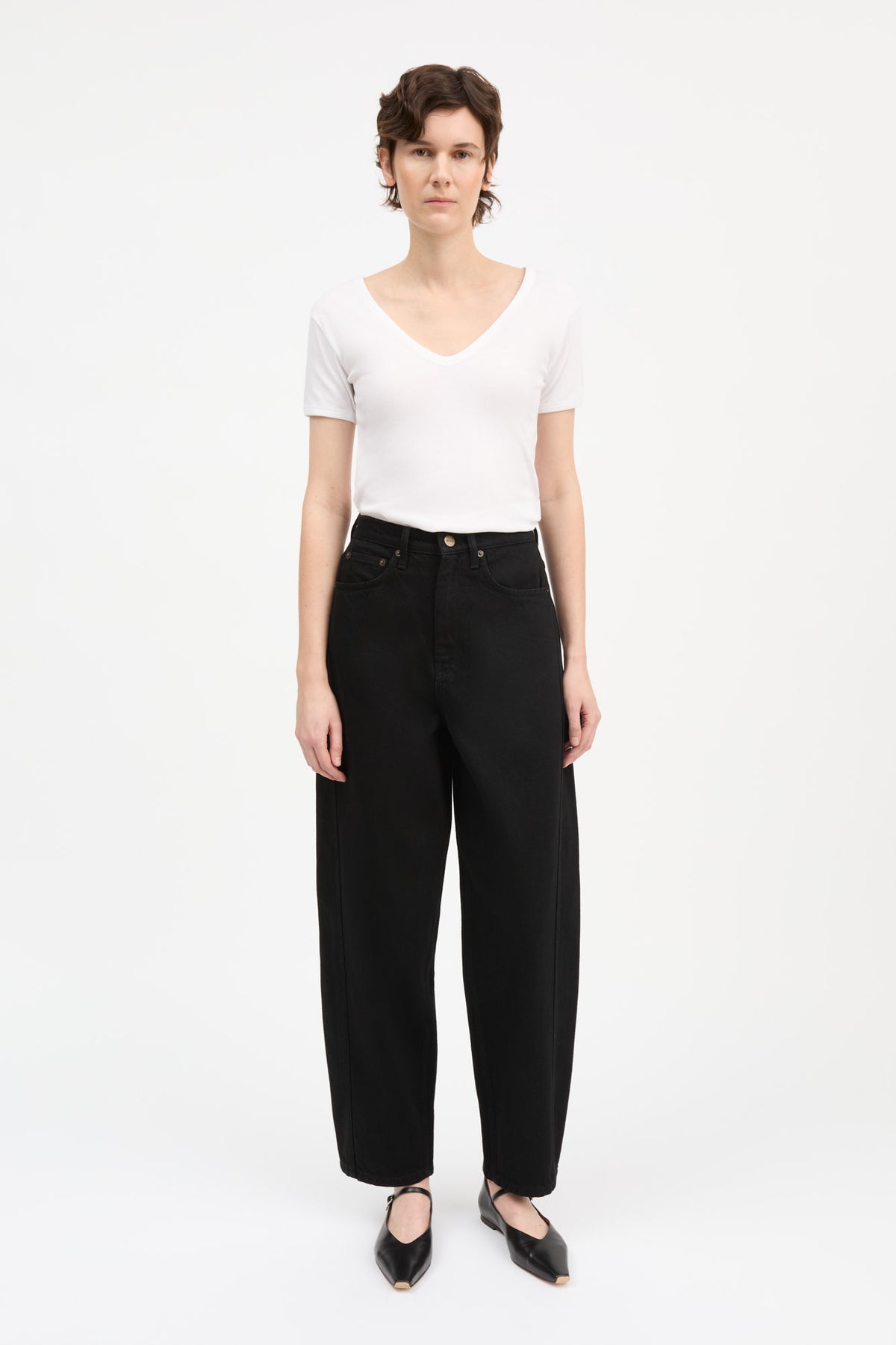 A model stands against a plain white studio background wearing a white short-sleeve V-neck top, black wide-leg cropped pants, and black pointed flats.