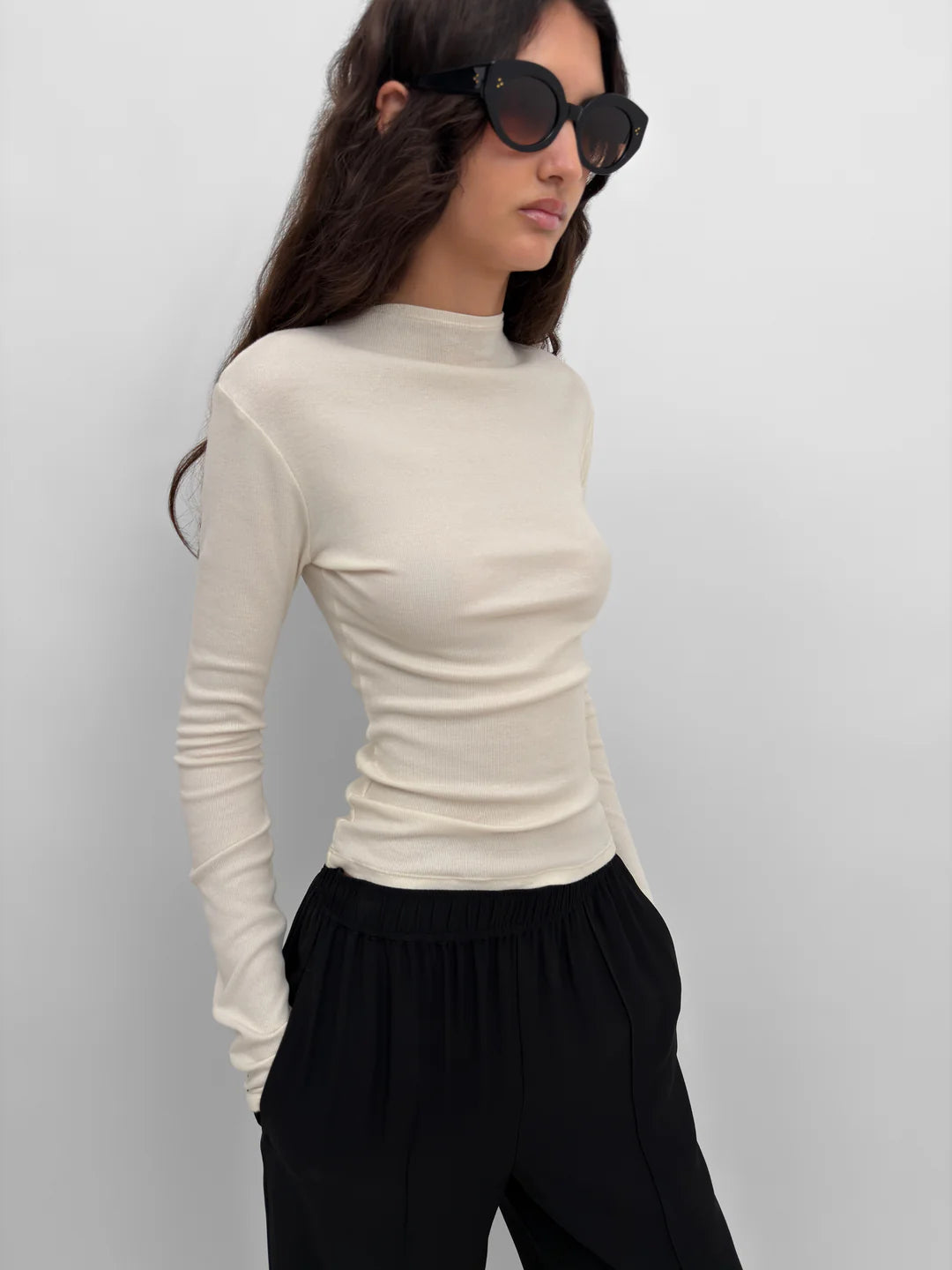 Woman wearing a beige long-sleeve top and black pants against a light gray background