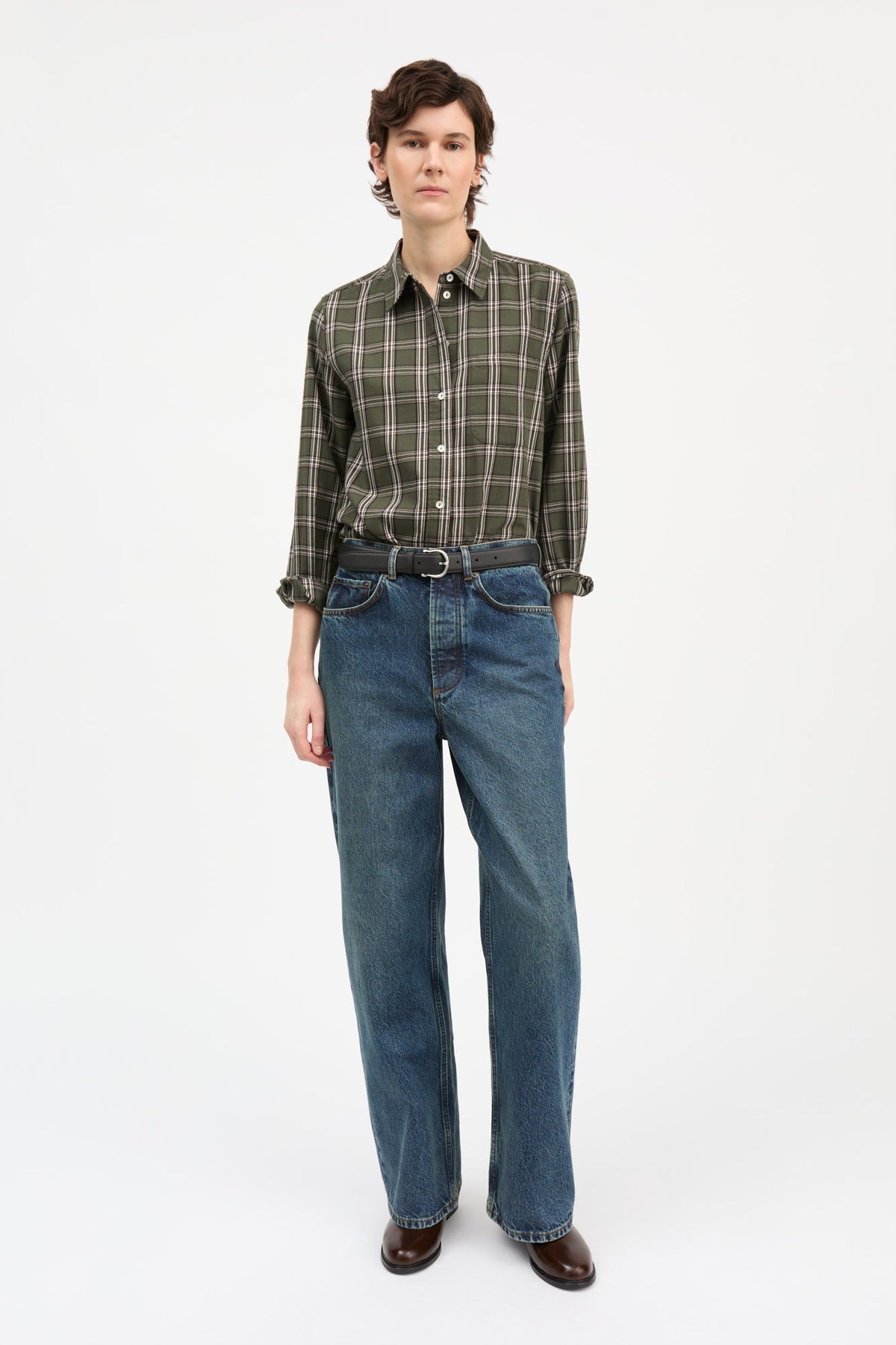 A model stands against a plain white studio background wearing a long-sleeve green and brown plaid button-down shirt, blue jeans, a dark belt, and brown leather shoes.