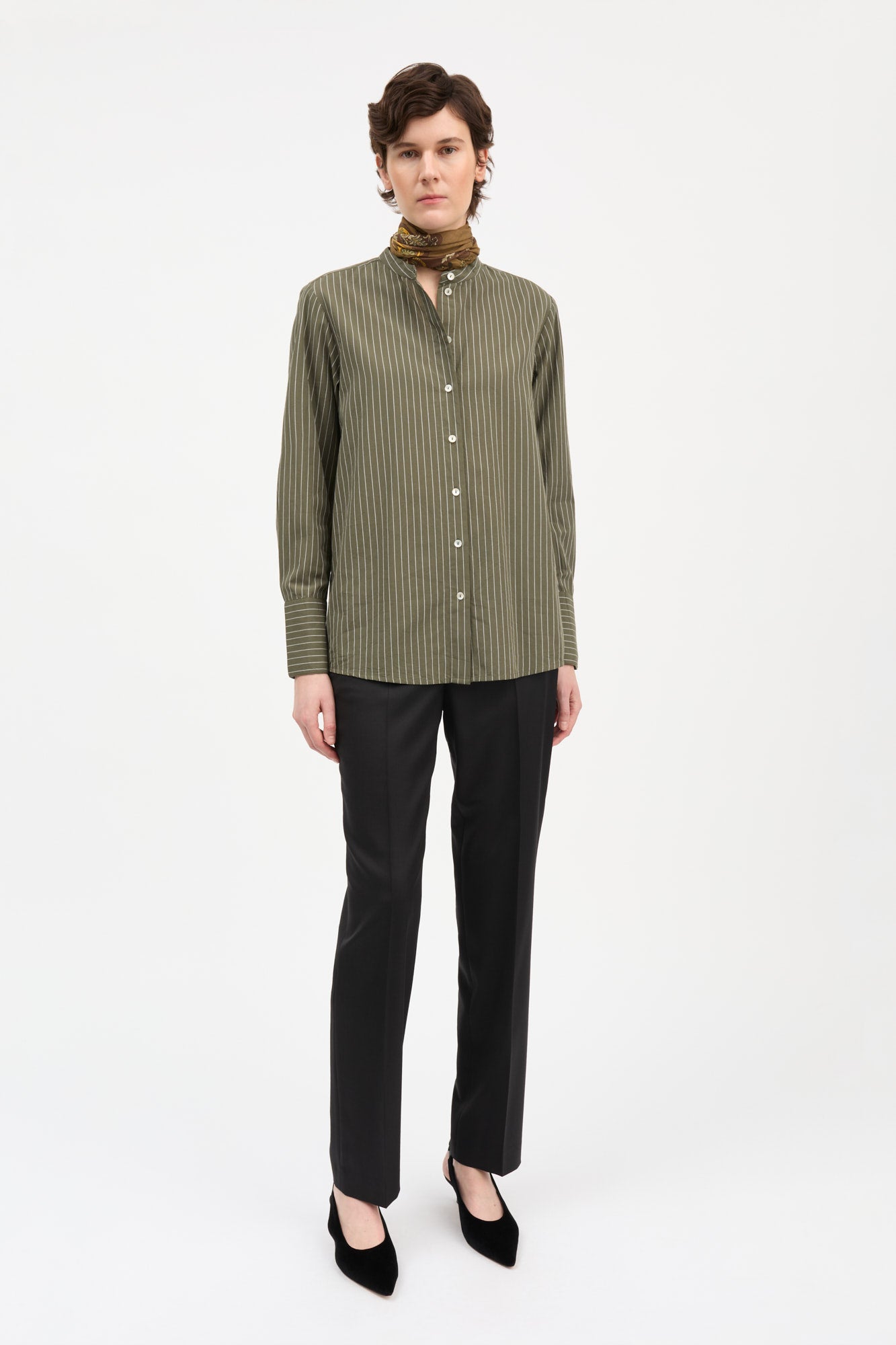 A model stands against a plain white studio background wearing a long-sleeve button-front shirt with vertical olive green and beige pinstripes, black trousers, black pointed shoes, and a brown patterned scarf.