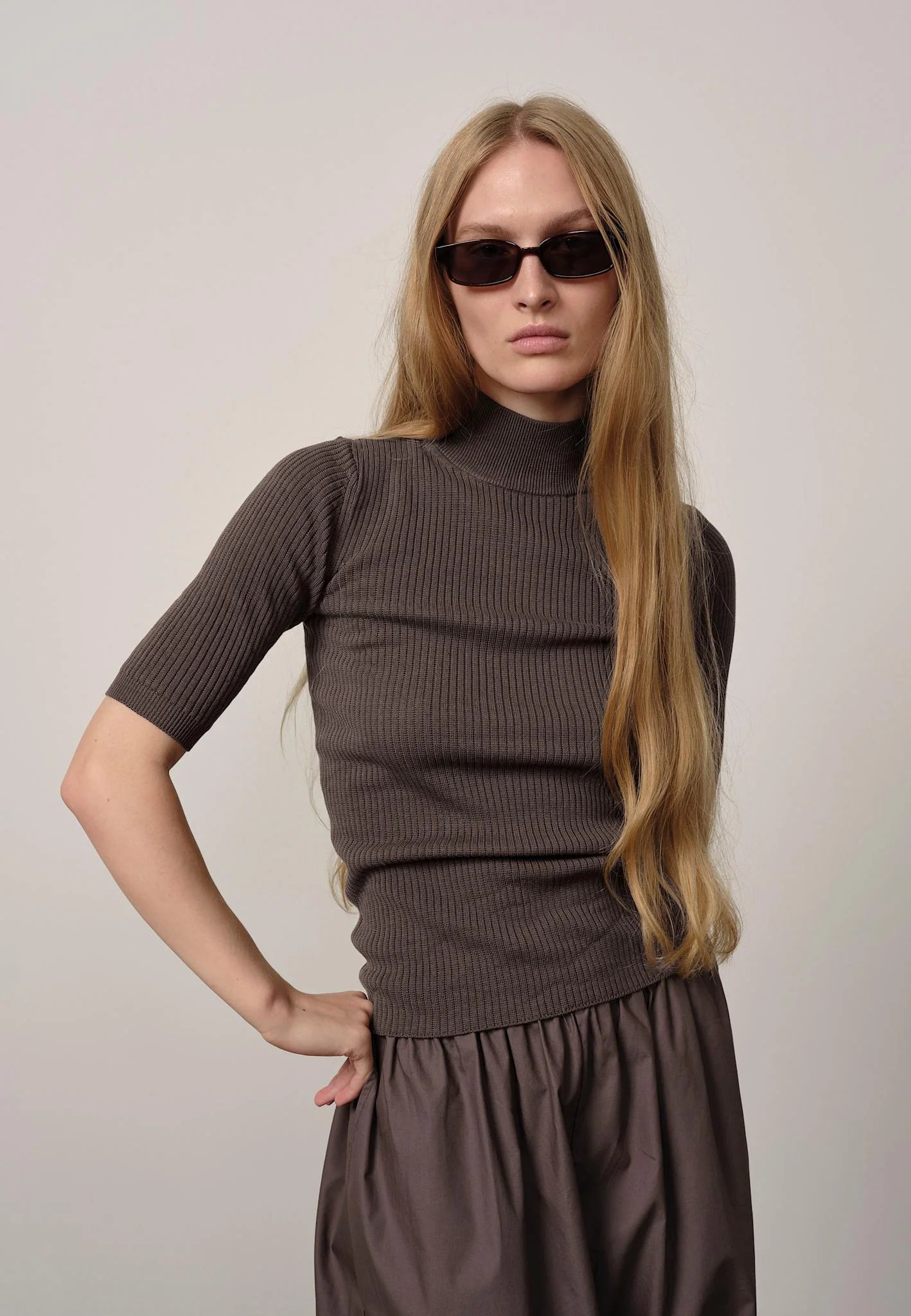 Woman wearing a brown ribbed sweater and skirt with sunglasses on a plain background