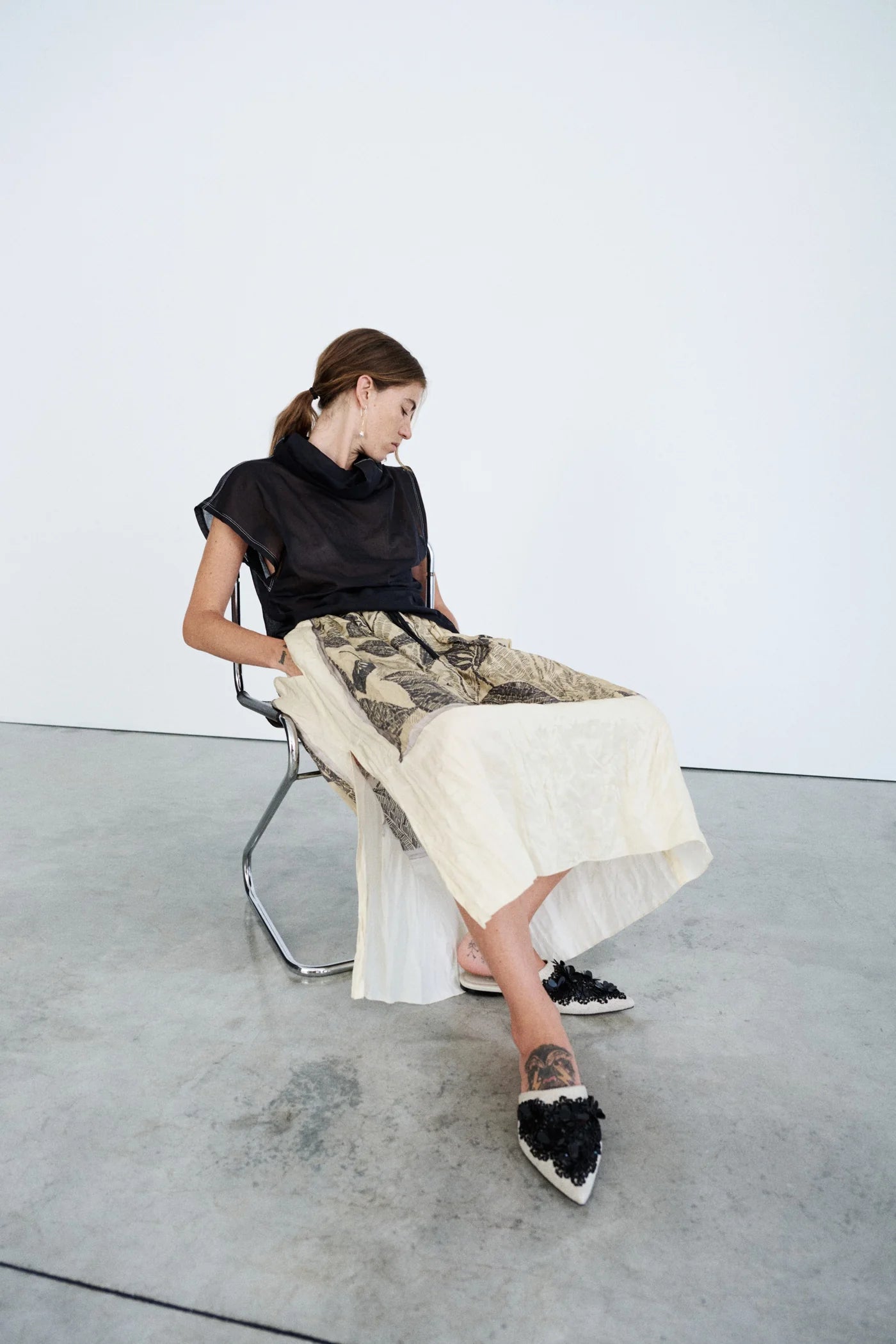Woman sitting on a chair wearing a black top and patterned skirt with white shoes.