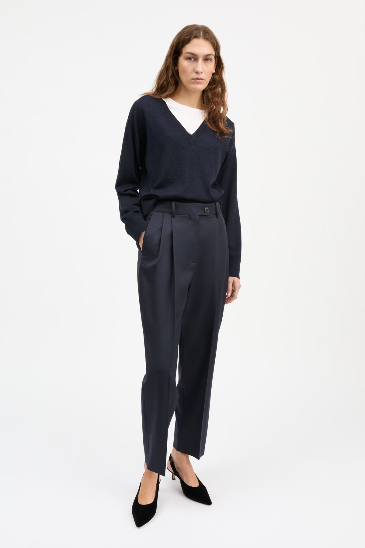 A woman modeling a dark navy blue long-sleeve V-neck top and matching high-waisted navy trousers with black pointed-toe heels, shot against a plain white studio background.