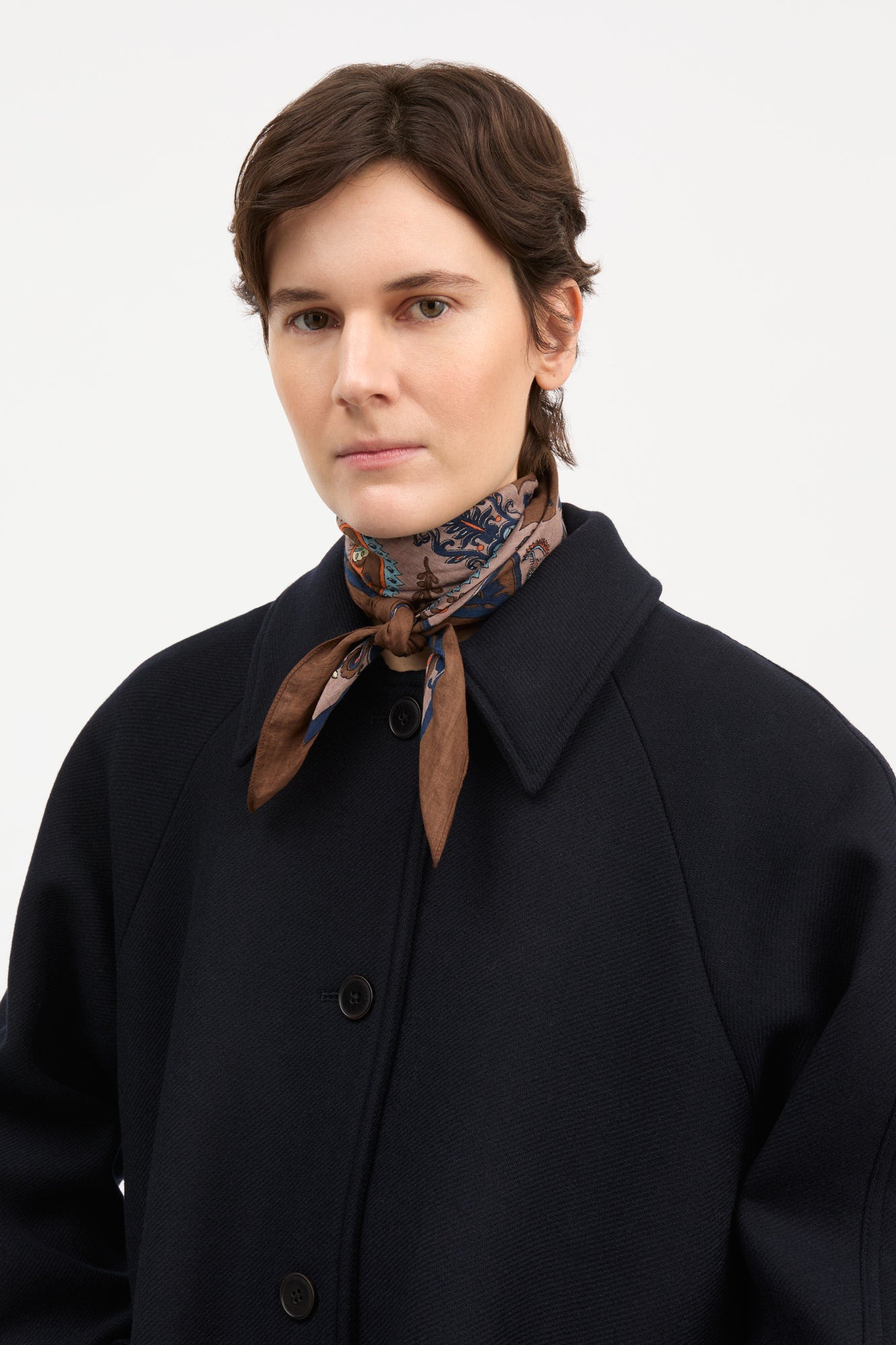 A model wearing a dark navy wool coat, a brown patterned silk neck scarf, and a black leather belt, photographed against a solid light gray background.