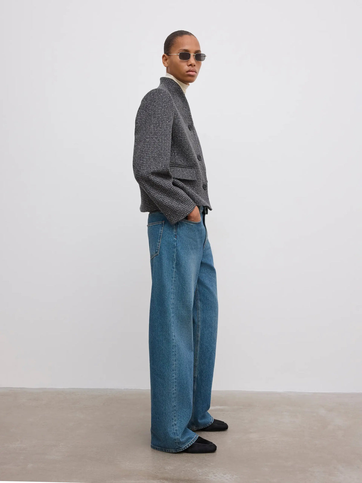 A model stands in profile wearing a gray textured jacket, wide-leg blue jeans, black shoes, and sunglasses against a plain light gray studio wall and concrete floor.