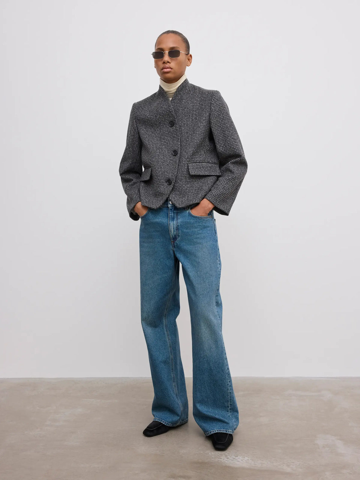 A model wearing a gray textured cropped jacket, wide-leg blue jeans, a cream turtleneck, and black shoes, standing against a minimalist light gray studio background.