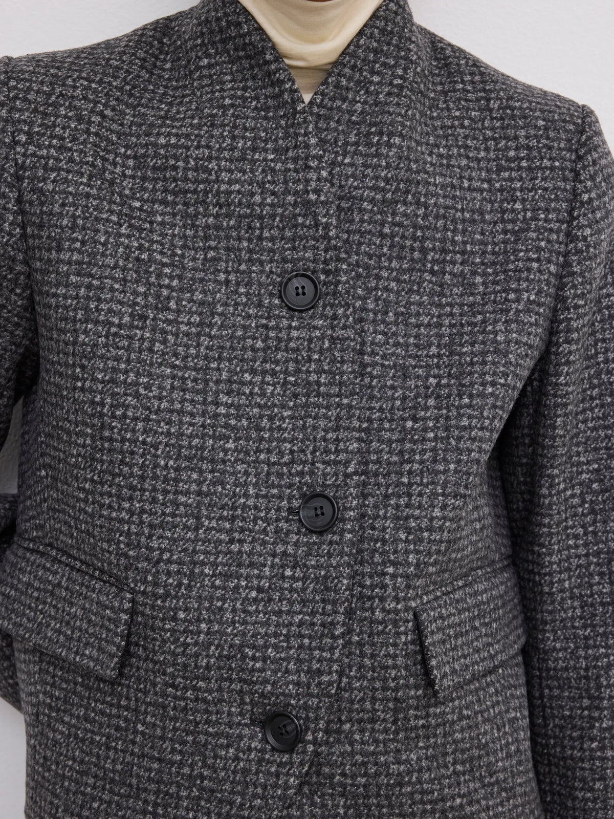 Close-up of a dark gray and black textured tweed jacket with a mandarin collar, three black buttons, and flap pockets, worn over a cream turtleneck.