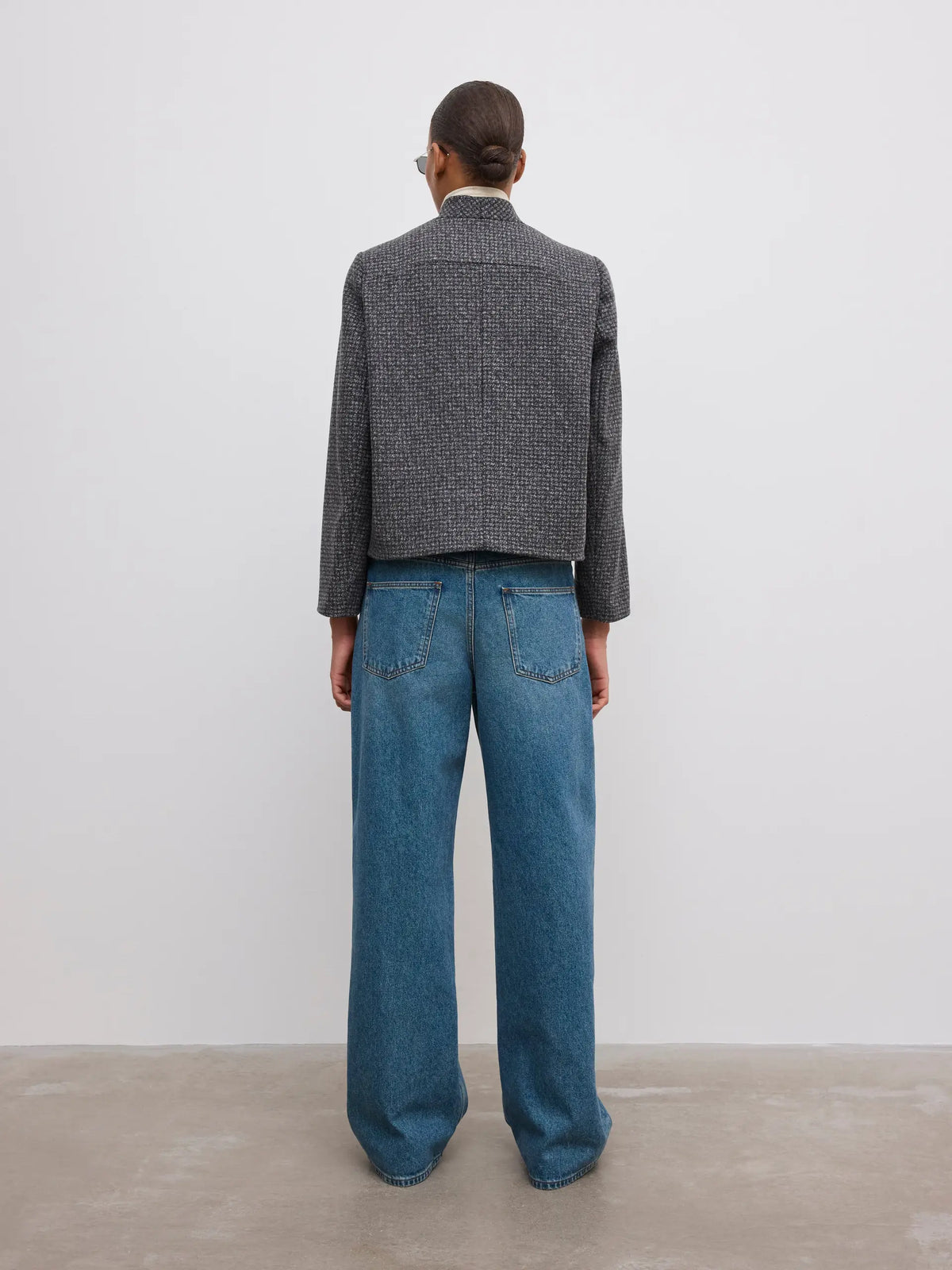 A model viewed from the back wearing a dark gray textured cropped jacket and wide-leg blue jeans, standing against a minimalist off-white wall and concrete floor.