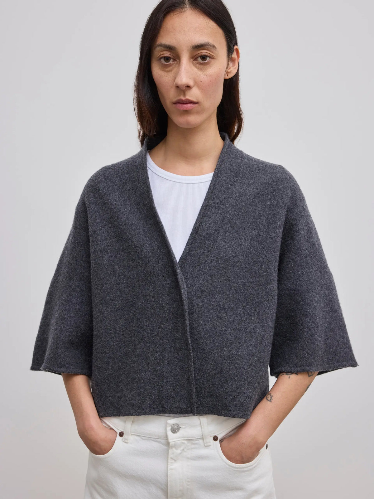 A woman wearing a dark gray open-front cropped cardigan with wide three-quarter sleeves over a white t-shirt and white jeans, standing against a light gray studio background.