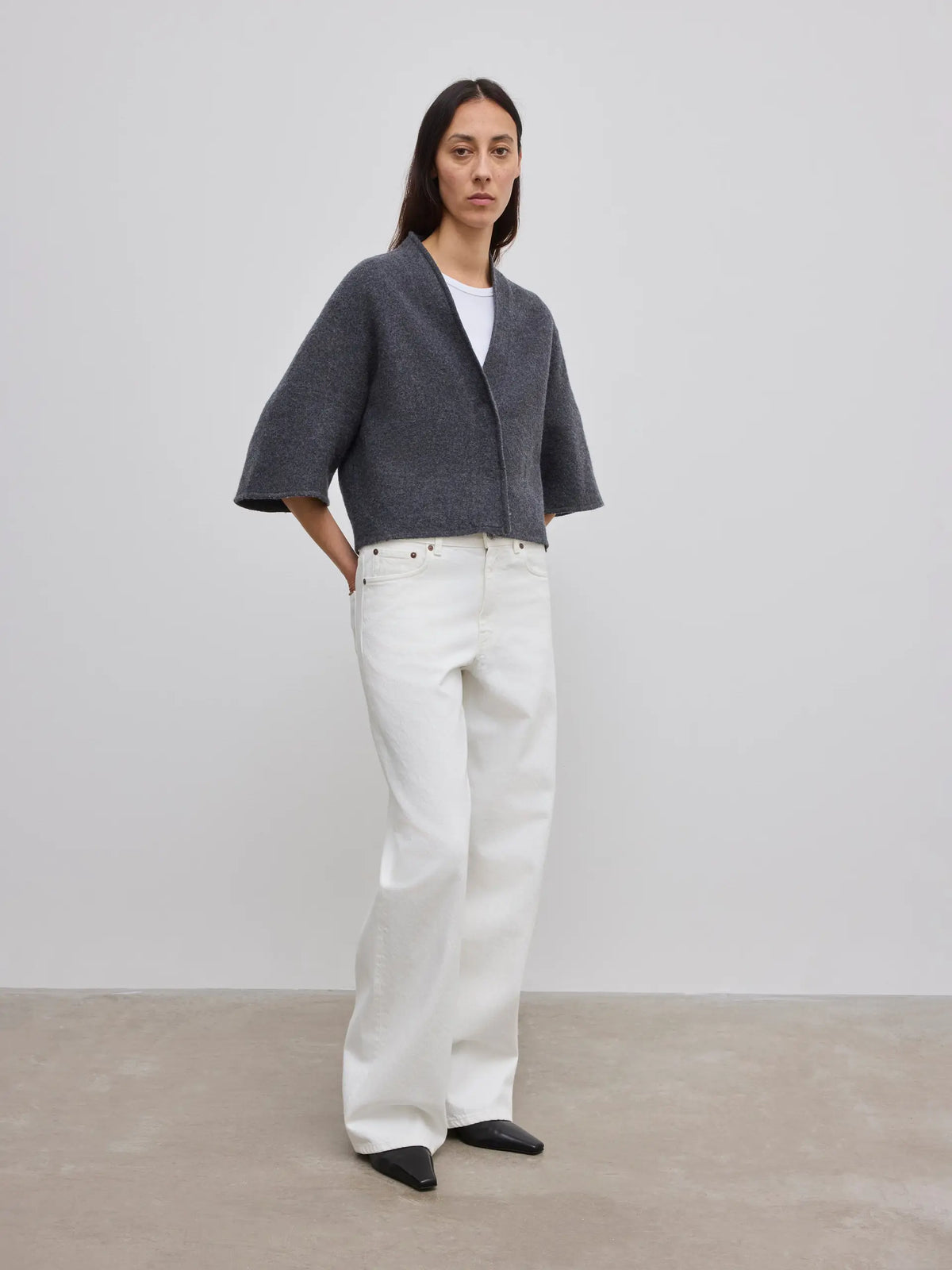A model stands in a minimalist studio setting wearing a dark gray cropped cardigan with wide three-quarter sleeves, white wide-leg trousers, and black pointed shoes.