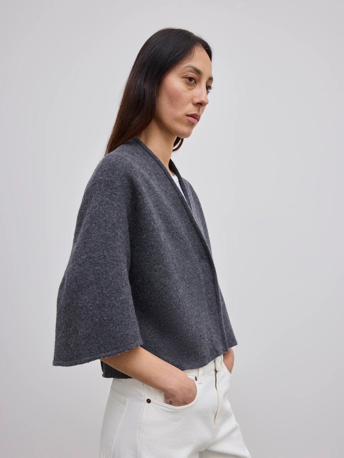 A woman wearing a dark gray cropped bolero jacket with wide, draped sleeves and white pants, photographed against a neutral light gray background.