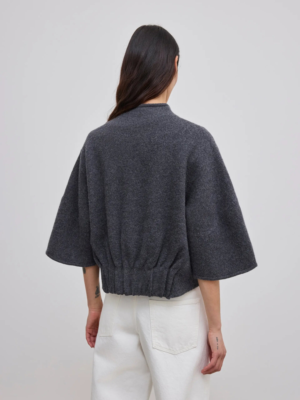 A woman shown from the back wearing a dark gray cropped knit cardigan with wide three-quarter sleeves and white pants, against a plain light gray studio background.