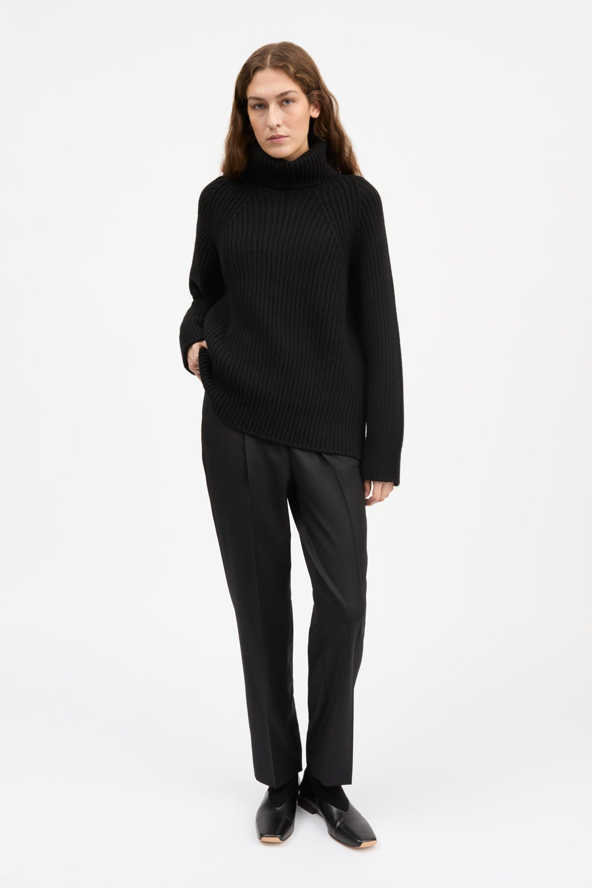 A woman modeling a black ribbed turtleneck sweater and black trousers against a plain white studio background.