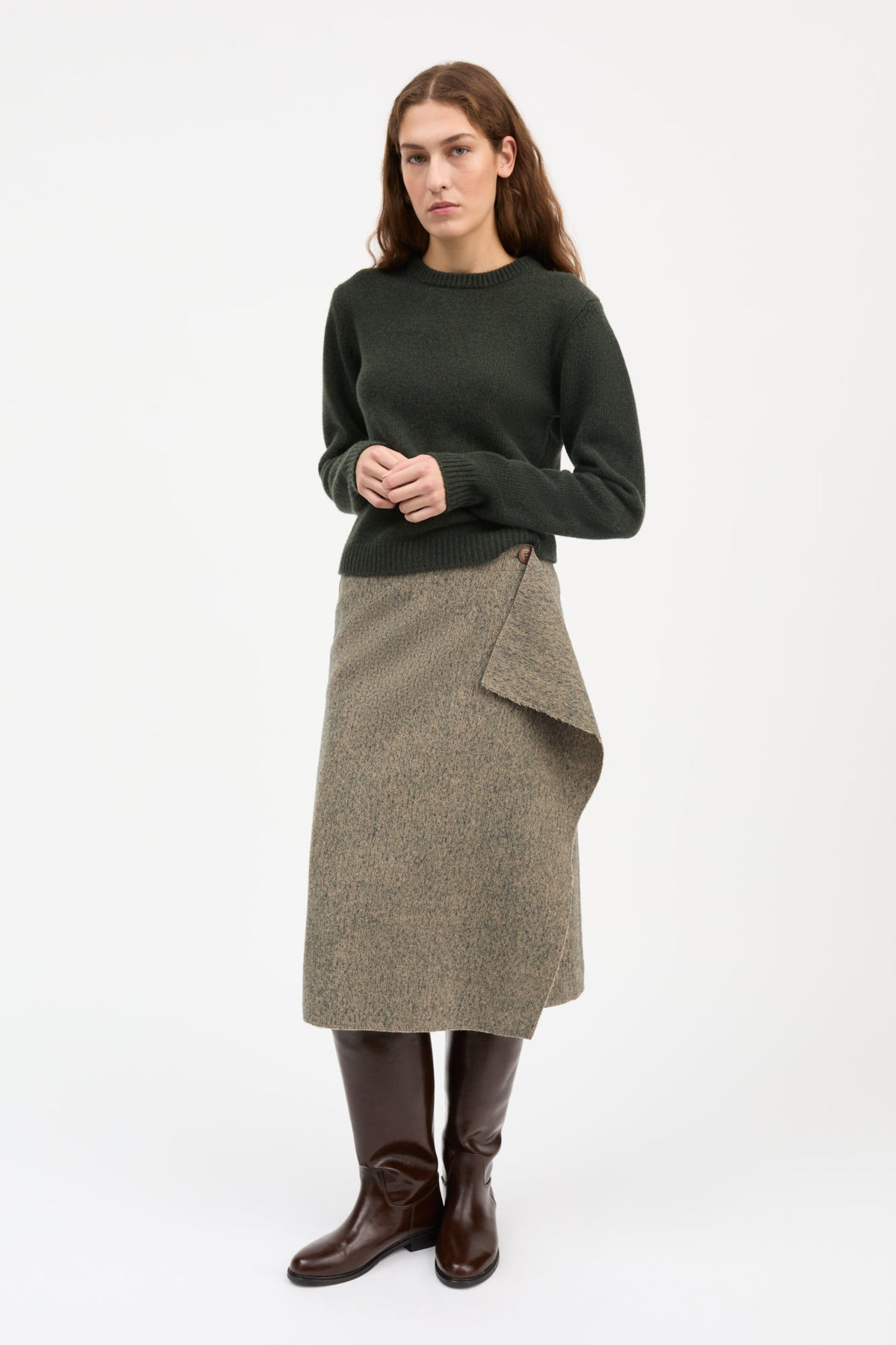 A woman stands against a plain light gray studio background wearing a dark green long-sleeve knit sweater, a textured taupe wrap-style midi skirt, and dark brown knee-high boots.